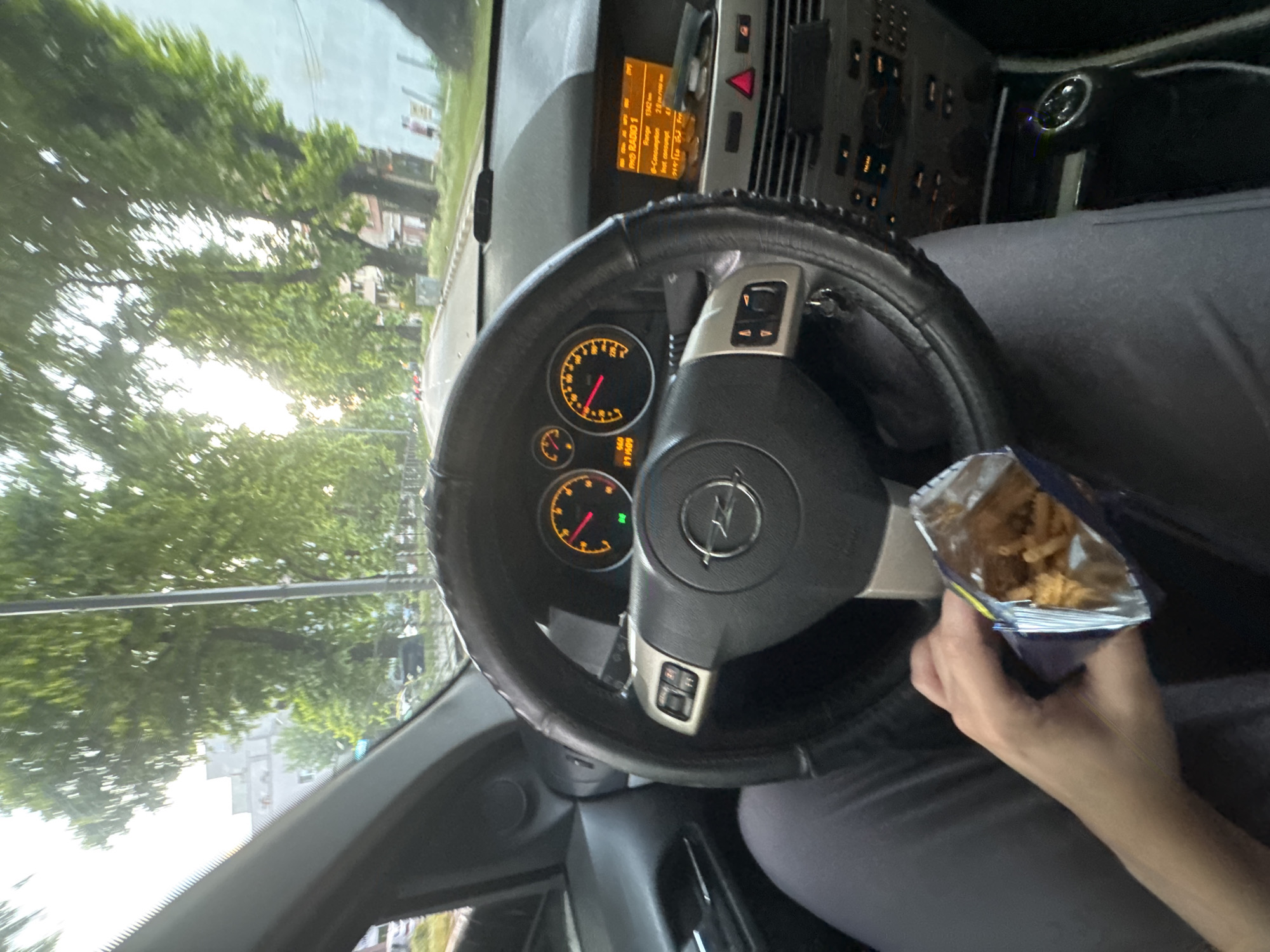 The image depicts the interior of a car, specifically an Opel model, from the driver's perspective, looking forward through the windshield onto an urban street in Asenovgrad, Bulgaria. The driver's left hand is visible in the lower center, holding an open blue and silver foil snack bag containing light brown, irregularly shaped snacks, likely consumed while the vehicle is either stationary or moving at a low speed.
The car's dashboard features a steering wheel with a black, textured cover and a silver Opel emblem at its center. Multi-function controls are present on both horizontal spokes of the steering wheel. The instrument cluster shows two primary gauges illuminated with an orange backlight: a tachometer on the left with its needle around 1000-2000 RPM, and a speedometer on the right with its red needle indicating a speed of approximately 20-30 units (likely km/h). Below the speedometer, a green indicator light is illuminated. The odometer displays "91909" and a trip meter "660". The center console includes an infotainment screen displaying "RMO RADIO 1," along with legible text fragments such as "Range 1542 km" and "S-Consumption 2.6 l/100km." Below the screen are air vents and various control buttons, including a red triangular hazard light switch. The driver's legs, clad in dark pants, are also visible.
Through the windshield, a tree-lined street is visible under bright, possibly overcast, daytime conditions. Mature trees with dense green foliage flank both sides of the road, which appears to be paved with asphalt and has a visible curb. In the distance, other vehicles, including some red-colored cars, are present on the road. Buildings are discernible behind the trees; on the left, a multi-story building, and on the right, a lighter-colored building with what appears to be a red figure on a sign. The overall environment suggests a typical urban setting with residential or commercial structures common in a city like Asenovgrad.