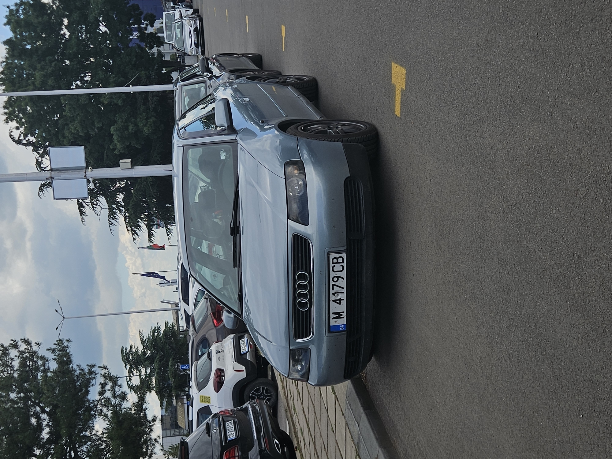 The image shows a parked light blue Audi A3 with a Bulgarian license plate "BG M 4179 CB" on a paved surface. The car is in the foreground, taking up a significant portion of the image. The background displays a parking lot with several other vehicles, trees, lampposts, and buildings. The sky is cloudy. The license plate on the Audi suggests it is registered in Sofia, Bulgaria, as the letter "M" typically denotes Sofia for Bulgarian plates, and "BG" indicates the country code.