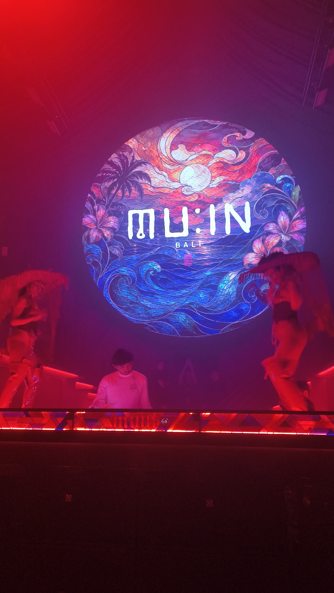 The image captures a nightclub scene in North Kuta, Indonesia, featuring a large circular graphic projected onto a screen behind a DJ booth. The graphic depicts a stylized sunset over a tropical ocean with palm trees and vibrant floral elements. The text "MU:IN BALI" is prominently displayed in white lettering within the graphic.
In the foreground, a DJ wearing a light-colored long-sleeved shirt stands behind a DJ console adorned with red LED lighting. To the left and right of the DJ, two performers, likely dancers, are visible. The performer on the left wears a light-colored, elaborate costume with feather headdresses and shimmering legwear. The performer on the right is also adorned in a similarly elaborate costume. The overall lighting is dominated by red and pink hues, creating a club atmosphere. The background is dark, with some additional figures and lighting visible in the distance, suggesting a crowded venue. The scene conveys a festive and energetic ambiance typical of a nightclub or party event.