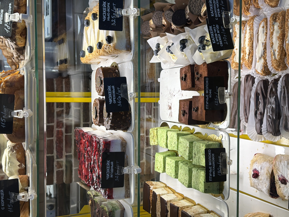 This image captures a brightly lit, multi-tiered retail display case filled with a variety of freshly prepared baked goods and desserts, located in a commercial establishment within посёлок 1-го отделения совхоза "Масловский", Russia.
The display features at least four glass shelves supported by yellow vertical beams. The background exhibits a reflection of a red brick wall. On the top shelf, various sweet rolls are arranged, including cinnamon rolls ("С КОРИЦЕЙ") priced at 115 rubles per piece, and other cream-filled pastries ("КРЕМОМ").
The second shelf from the top presents three distinct cheesecake varieties: a large rectangular "ЧИЗКЕЙК ЯГОДНЫЙ" (Berry Cheesecake) with a vibrant red berry topping, priced at 325 rubles per piece; several smaller, individual "ЧИЗКЕЙК ОРЕХОВЫЙ" (Nut Cheesecake) with dark chocolate coating and golden embellishments, priced at 360 rubles per piece; and a large rectangular "ЧИЗКЕЙК НЬЮ-ЙОРК" (New York Cheesecake) garnished with blueberries, priced at 325 rubles per piece. All items are presented on white rectangular plates.
The third shelf features a selection of layered cakes and individual desserts. On the left are slices of layered cakes with dark tops. Adjacent to them are rows of small, rectangular "ПТИЧЬЕ МОЛОКО ФИСТАШКОВОЕ" (Ptyche Moloko Pistachio) cakes, colored green and priced at 130 rubles per piece. To their right are similar "ПТИЧЬЕ МОЛОКО КОФЕ" (Ptyche Moloko Coffee) cakes, dark brown with two coffee beans on top, priced at 125 rubles per piece. Further right, white cupcakes topped with cream and blueberries ("КАПКЕЙК С КРЕМ-ЧИЗ СВЕЖЕЙ ЯГОД") are displayed in paper wrappers, priced at 210 rubles per piece, alongside other cupcakes topped with Oreo cookies ("КАПКЕЙК С ШОКОЛАДНОЙ КРЕМ-ЧИЗ").
The bottom shelf showcases pastries with fruit filling, powdered sugar, and two types of éclairs: one row of dark chocolate-coated éclairs and another row of lighter-colored, cream-filled éclairs. Each item or group of items is accompanied by a black price tag with white Russian text and a price in rubles, affixed with clear plastic clips. No individuals are visible in the frame.