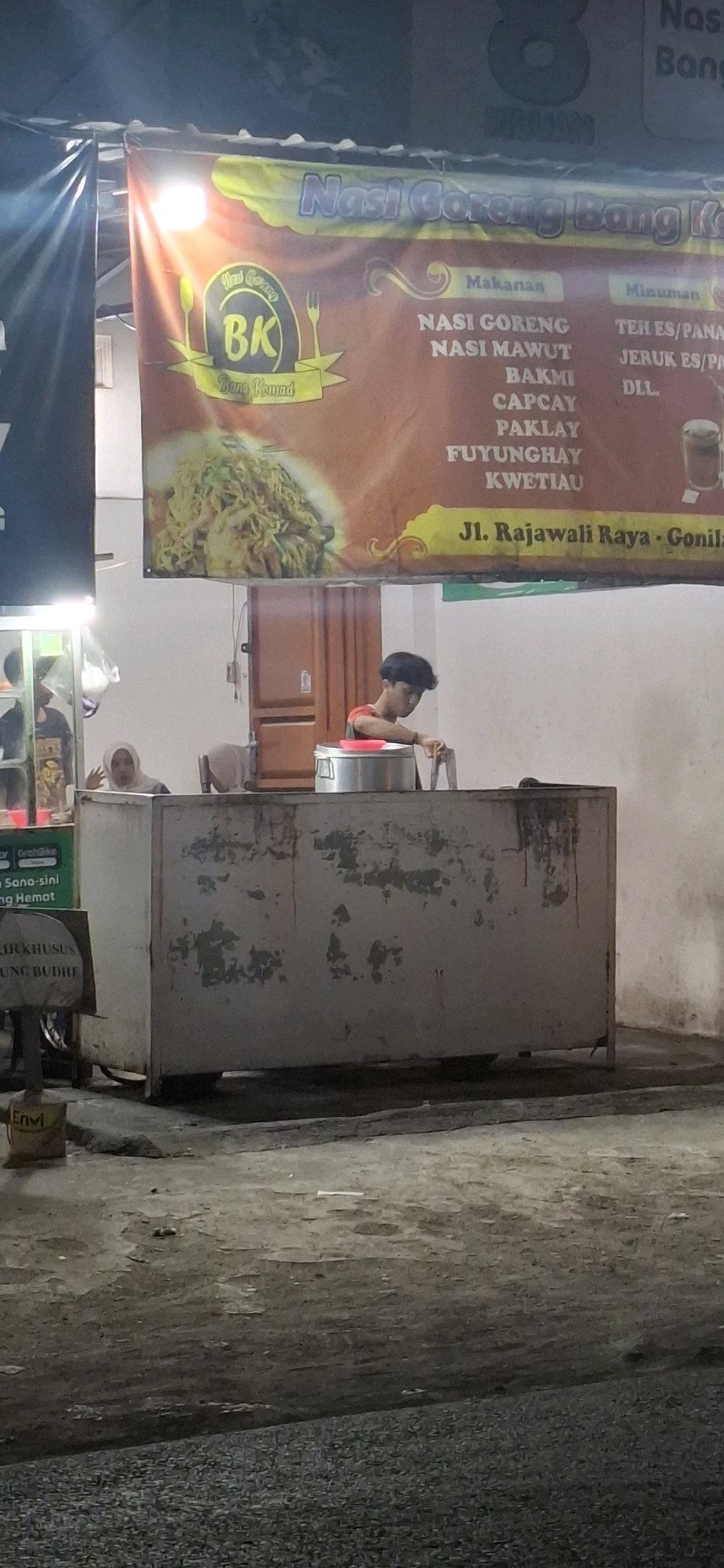 The image shows a night scene at a street food stall in Pabelan, Indonesia. The stall has a large banner advertising "Nasi Goreng Bang Komad" and lists various food and drink options such as "Nasi Goreng," "Nasi Mawut," "Bakmi," "Capcay," "Paklay," "Fuyunghay," "Kwietiau," "Teh ES/Panas," and "Jeruk ES/Panas." The banner also displays the address "Jl. Rajawali Raya - Gonila."
In the foreground, a young man with dark hair is seen working behind a metal food cart. He is wearing a red shirt and appears to be preparing food. To the left of the food cart, a woman wearing a hijab is visible, seemingly interacting with someone. The environment appears to be an outdoor setting with a paved ground. There are some signs and plastic bags visible, suggesting a typical street food vendor setup. The lighting indicates it is nighttime, with artificial lights illuminating the area.
