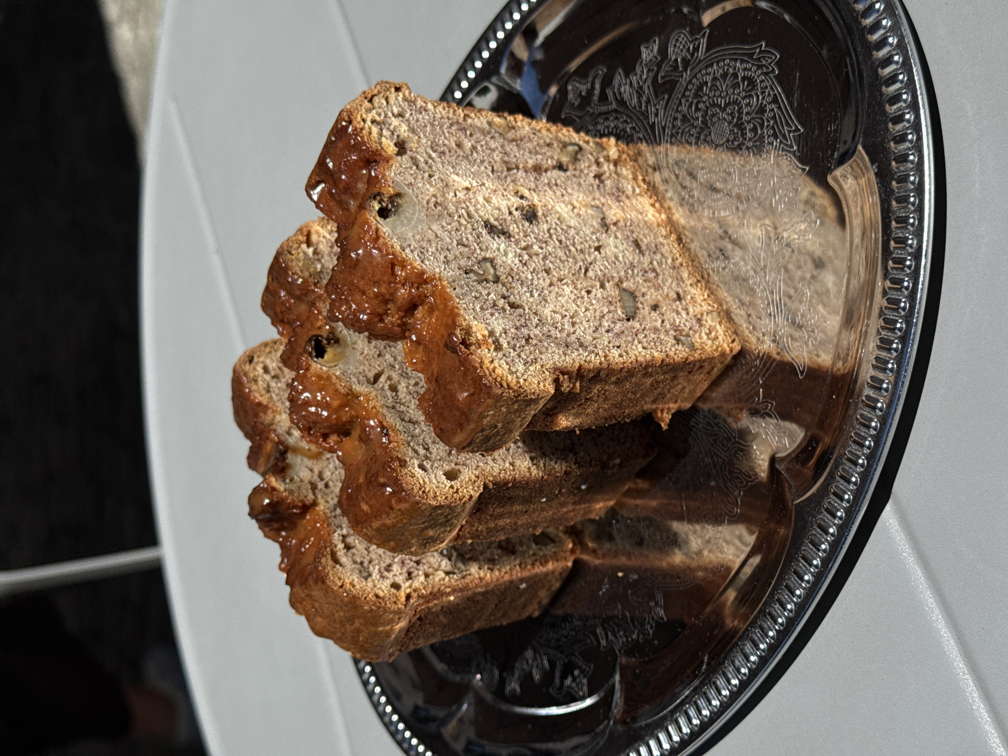 Three slices of banana bread are displayed on a silver platter with an ornate embossed design. The banana bread has a rich, brown hue and appears to be moist, with visible chunks of walnuts throughout. The top of the bread is glazed with a glossy, amber-colored syrup. The platter rests on a light-colored surface, possibly a table, which is set against a blurred background that suggests an outdoor setting with dappled sunlight. The scene is captured in Vracar Urban Municipality, Serbia, possibly on a bright, sunny day. There are no people clearly visible in the image.