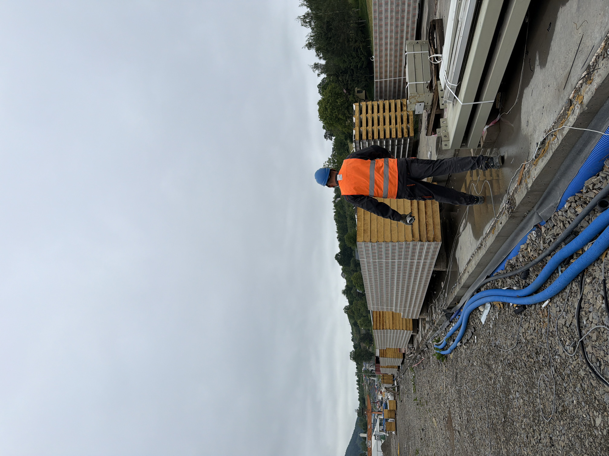 A construction worker wearing a blue hard hat and an orange reflective vest walks away from the camera on a construction site in Męcina, Poland. He is wearing black pants with red accents and dark work boots. In front of him, stacked in neat rows, are large panels, likely for building construction, with yellow insulation cores and striped outer surfaces. To the left, more stacks of panels are visible, as well as construction materials and equipment. Blue corrugated pipes and black electrical cables are laid out on the gravelly ground. In the background, trees and hills suggest a rural or semi-rural setting. The sky is overcast.