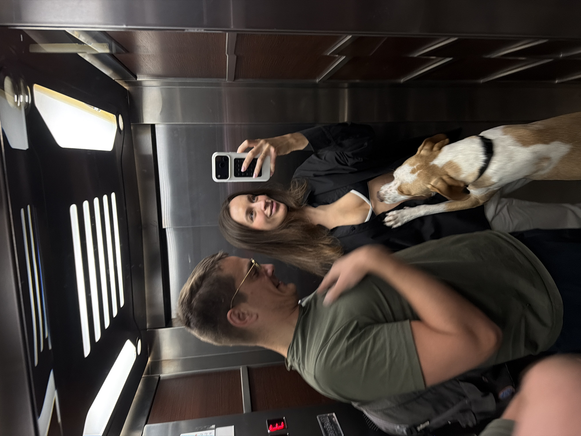 The image depicts two adults and a dog in an elevator. The female adult is holding a smartphone and taking a selfie. The male adult is standing next to her, smiling. The dog is positioned between them. The elevator's interior features a metal and wood design with illuminated panels. It is not possible to determine the location of the image in the city of Budva, Montenegro without further information or context.
