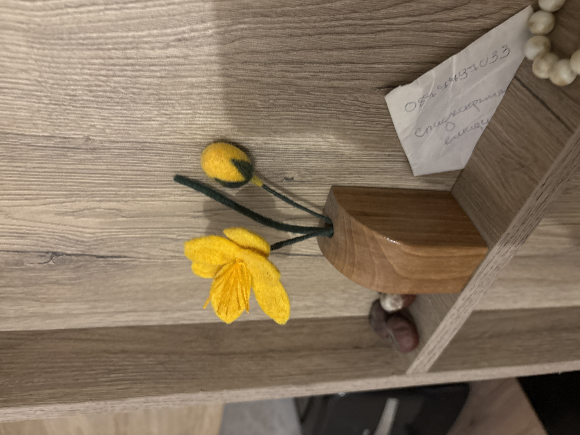 The image shows a close-up of objects on a wooden shelf. A small, felt yellow flower and bud are in a wooden vase. A piece of paper with handwriting on it is next to the vase. The text on the paper includes a phone number beginning "0894" and the words "Специална единц". There are some white beads to the right of the paper. Brown objects are visible below the vase. The location is not identifiable from the image.