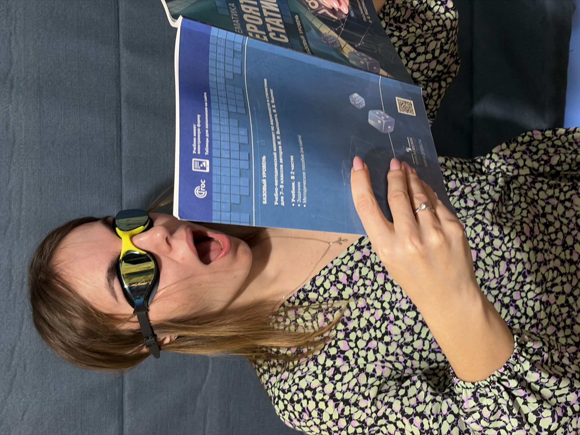 A woman is pictured indoors, possibly lying on a couch or bed, given the texture of the grey surface behind her. She is wearing black and yellow swimming goggles on her head and a light green dress with small black and purple patterns. She has her mouth open as if in surprise or excitement. In her hands, she holds open a book titled "MATEMATIKA, VEROYAT, STATISTI" (Mathematics, Probability, Statistics) written in Russian, with the author's name partially visible: "V. Yaschenko". The book cover also contains Russian text indicating it is a basic level учебник (textbook). The logo "Croc" is visible on the book's cover. The image provides no information that places the subject in Izhevsk, Russia.