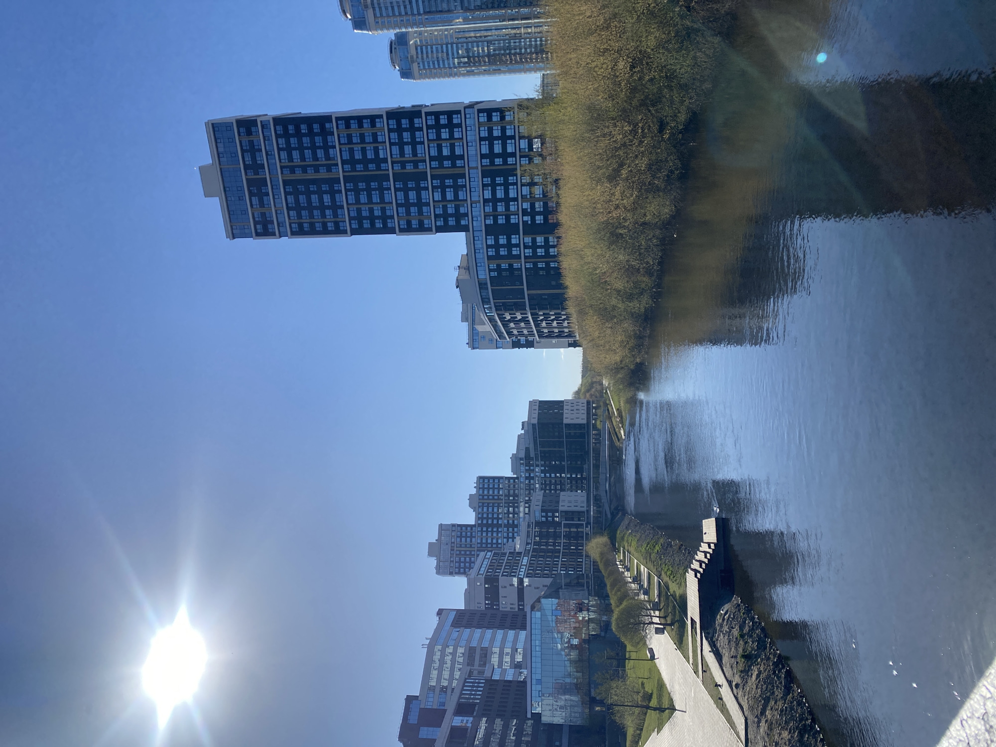 This media file presents a bright, sunny daytime scene of a modern urban landscape in Yekaterinburg, Russia, featuring a river and a collection of contemporary buildings.
The scene is dominated by a clear, vibrant blue sky, with the sun shining intensely in the lower-left corner, causing a noticeable lens flare. A wide river or canal runs vertically down the right side of the image, its calm surface reflecting the blue sky and distorted shapes of the surrounding buildings and trees.
Along the far bank of the river, a dense line of trees or bushes with nascent foliage creates a natural boundary. On the near bank, a paved walkway or promenade stretches along the water's edge, featuring neatly planted small trees and patches of green. A small dock or structure with steps extends into the water from this bank in the mid-ground.
The left side of the image is filled with several modern high-rise buildings. These structures boast varied architectural designs, mostly featuring dark facades with numerous rectangular windows, interspersed with lighter sections and extensive glass panels that reflect the sky and other buildings. Some buildings show stepped or curved designs, adding to the contemporary aesthetic.
No specific activities or individuals are visible, conveying a sense of quiet urban life. The clean lines of the architecture, the clear sky, and the calm water suggest a peaceful, well-maintained city environment. There is no visible text in the image.