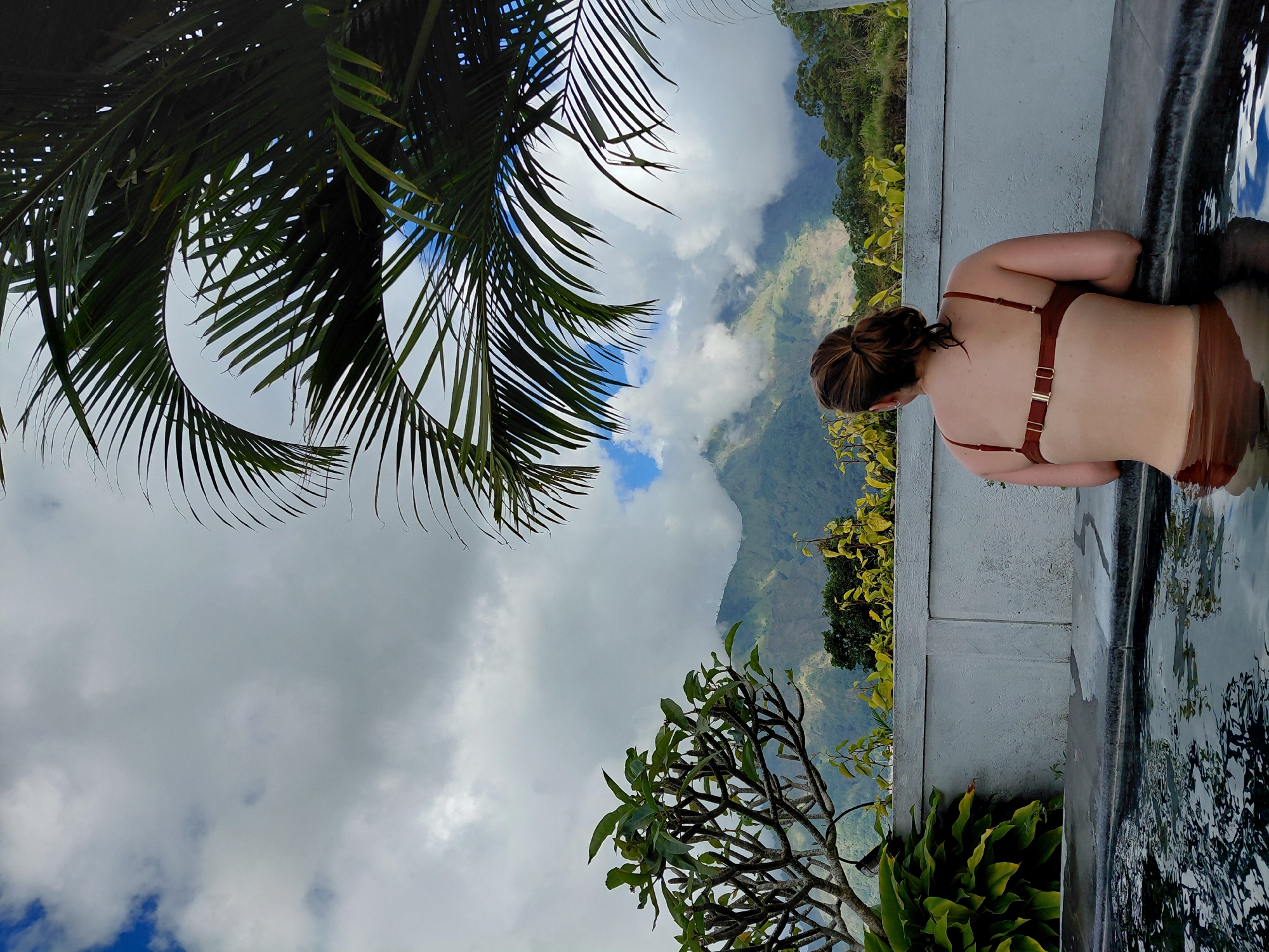A woman with brown hair pulled back in a bun is seen from behind, resting her arms on the edge of a dark-bottomed pool. She is wearing a brown bikini. The pool appears to be an infinity pool, with the water reflecting the sky and surrounding environment. In the background, a gray concrete wall with visible joints separates the pool area from a lush landscape. Beyond the wall, a frangipani tree with green leaves and thin branches is visible on the left. Further in the distance, a mountain range with some clouds obscuring the peaks is present. Palm fronds arch over the scene from the upper right. The sky is a mix of gray clouds and patches of blue sky with scattered white clouds. The overall scene suggests a tropical or mountainous location. This scene is likely located in the city of Kintamani, Indonesia, known for its stunning views of Mount Batur and Lake Batur, often from elevated viewpoints with pools or restaurants.
