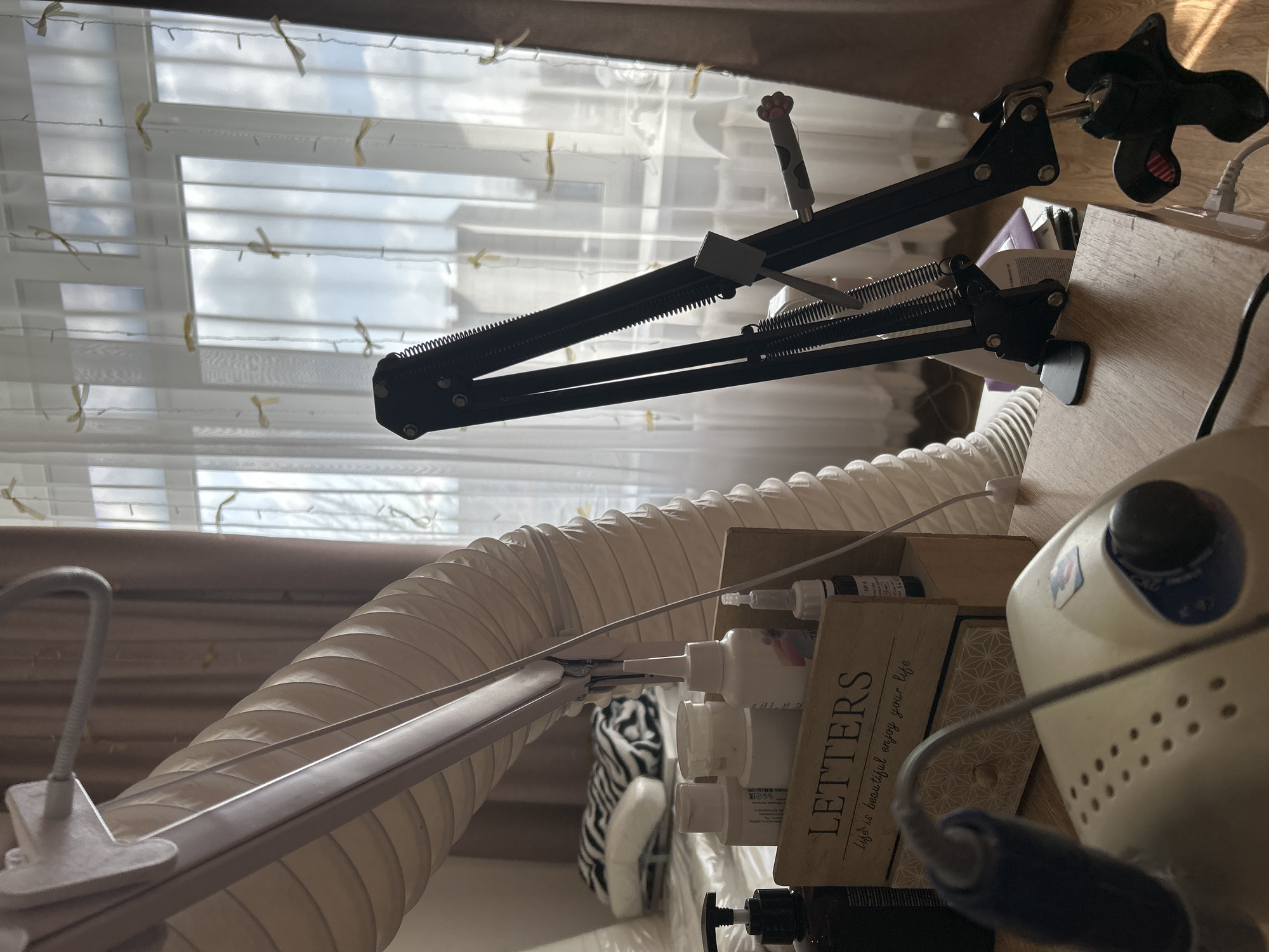 The image, likely taken indoors in a domestic setting in Chaikovsky, Russia, shows a portion of a workspace. The focal point is a black articulated arm, possibly a microphone stand, mounted on a wooden surface. It's positioned in front of a window covered by sheer curtains with decorative bows.
To the right, a portion of an electric nail file is visible. A storage box labeled "LETTERS" is filled with small bottles and other items.
A flexible, corrugated hose extends across the scene, suggesting a ventilation or extraction system. The presence of nail care tools and equipment indicates the space is likely used for manicures or similar beauty treatments. A striped pillow is visible in the background.
