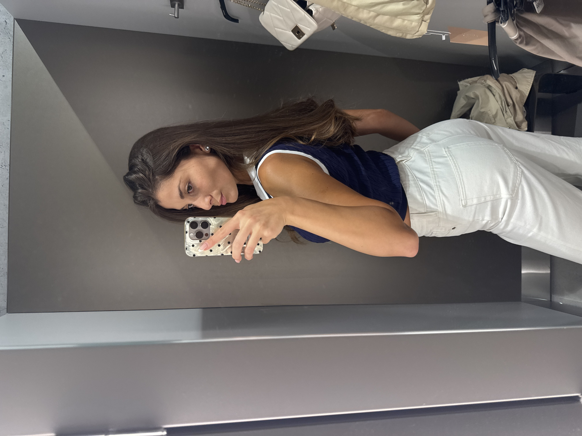 A woman is taking a selfie in a dressing room. She is wearing a navy and white tank top and white pants. She is holding a smartphone in her left hand. The dressing room has gray walls and a metal shelf at the bottom. There are clothes hanging in the background, including a white purse and a beige jacket. The location is not specified, so it is not possible to determine if it is in Burgas, Bulgaria.