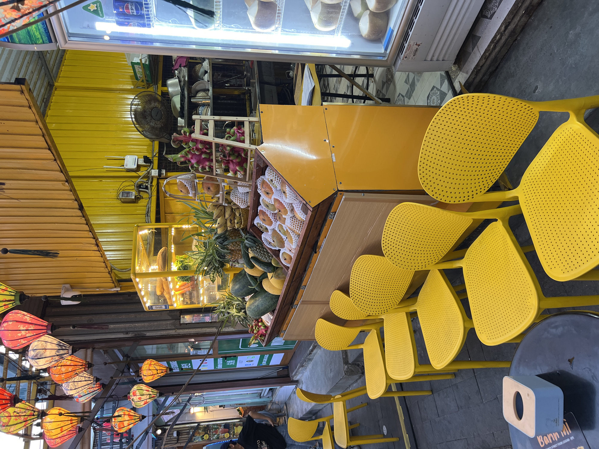A vibrant street scene unfolds with a focus on a fruit stall and seating area. In the foreground, several bright yellow chairs with perforated backs are arranged around dark tables, one of which holds a light blue tissue box. Behind the seating, a large, yellow display unit showcases an assortment of fruits, including watermelons, pineapples, dragon fruit, bananas, and other boxed fruits. The stall is illuminated by strings of colorful, illuminated lanterns hanging overhead. To the right, a white refrigerator displays coconuts on its shelves, with a Pepsi can visible on an upper shelf. The background reveals the exterior of a building with yellow corrugated metal walls, some electrical equipment, and what appears to be a fan. The overall impression is that of an outdoor market or food stall on a city street.