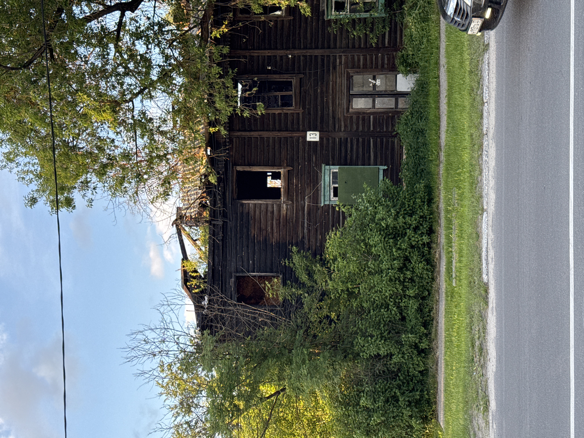 The image depicts an old, two-story wooden building appearing severely damaged, possibly from a fire, located beside a paved road. The building's exterior is made of dark, weathered vertical wooden planks, with several windows. Many windows are broken, missing glass, or boarded up; one on the lower level features a distinctive green frame and cover. A white plaque with the number "13" is clearly visible on the building's facade. The upper section and roofline show significant structural damage, with exposed, charred wood and a partially collapsed roof, indicating ruin or long-term neglect.
Lush green trees and bushes surround and partially obscure the building, particularly on the left side and in front. A dark cable or wire runs vertically down through the foliage on the left. To the right of the building, a grassy verge separates the property from a paved road, which features a solid white line along its edge. The front portion of a dark-colored vehicle, possibly a car or SUV, is visible on the road, facing away from the viewer. Its headlights appear illuminated or reflective, and a partial license plate is discernible but unreadable. The sky, visible through the trees and above the damaged structure, is blue with scattered white clouds, suggesting a daytime scene, possibly late afternoon given the soft lighting. No people or active events are depicted.