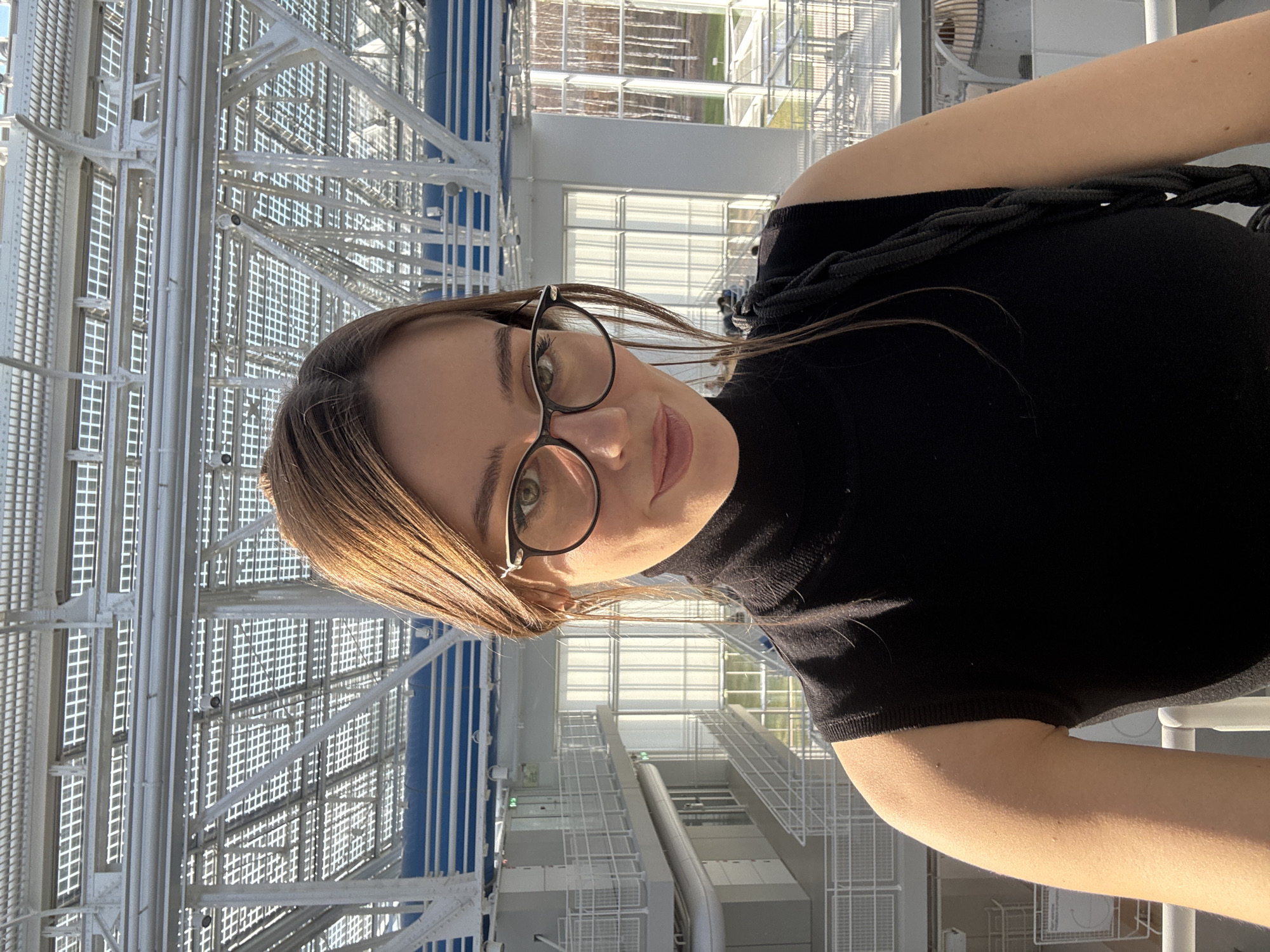 A young woman with brown hair and glasses is taking a selfie. She is wearing a black top and standing against a background of a modern architectural structure made of steel and glass. The structure appears to be a high viewpoint, offering a view of the surrounding city. Based on the architecture, it is likely the viewpoint at the Central Children's Store in Moscow, Russia. The light is bright, suggesting it is daytime.