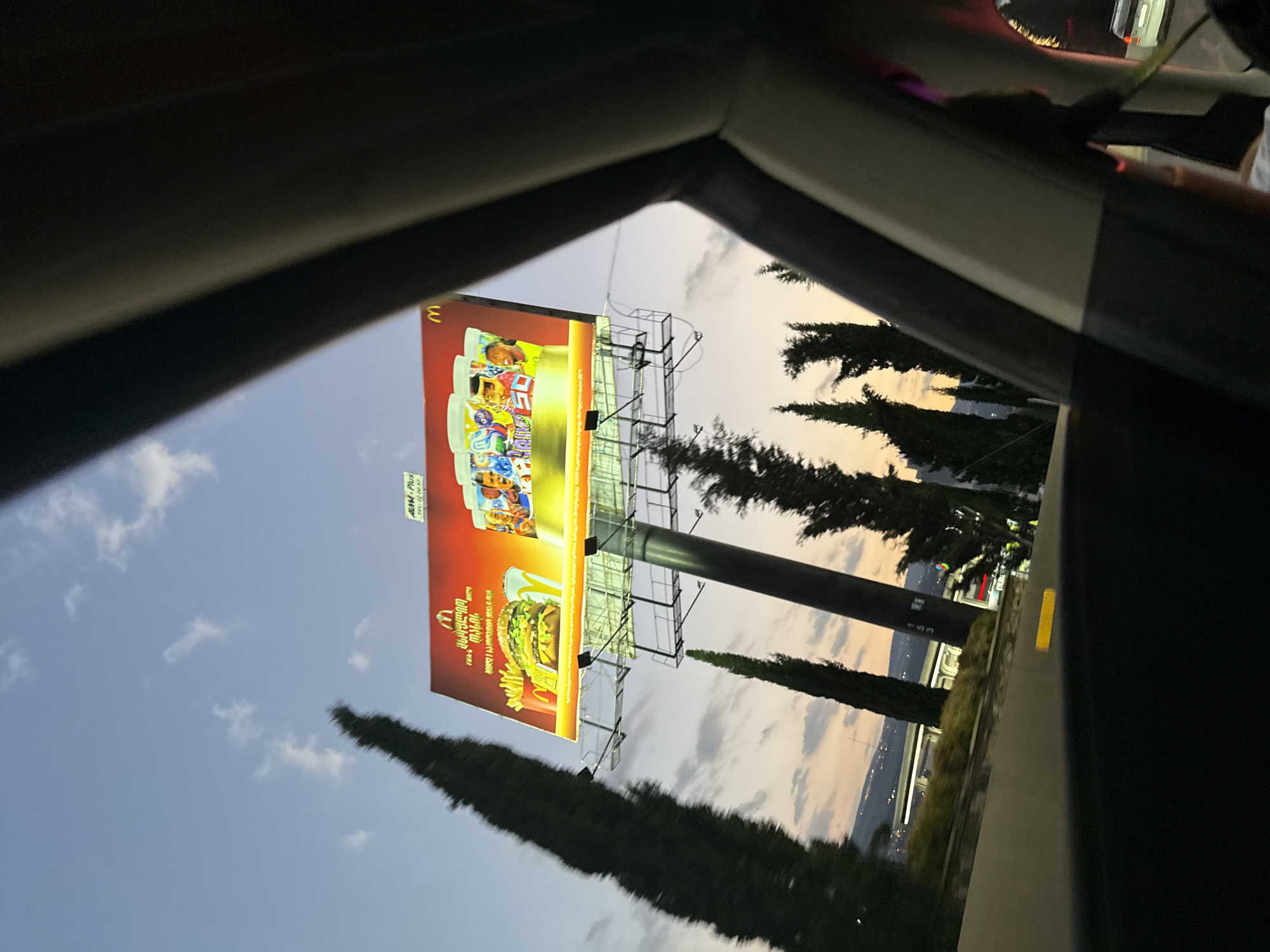 The image captures a billboard for McDonald's, displaying a Big Mac and drinks with images of soccer players. The billboard is illuminated and positioned along a road. Several tall, slender trees flank the billboard. In the background, a city skyline with lights suggests an urban environment, likely Tbilisi, Georgia, given the context of previous searches. The sky is a dusky blue with scattered clouds, indicating either dawn or dusk. The perspective is from inside a vehicle, with parts of the car's interior visible in the foreground.