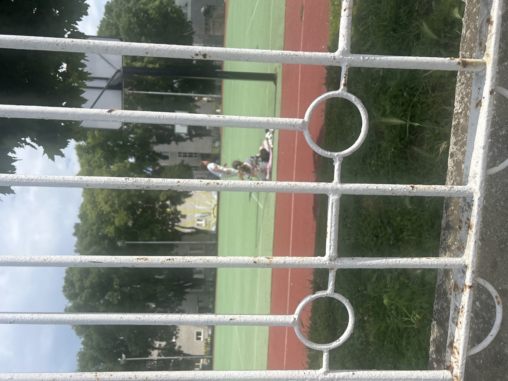 The image shows a view through a white, metal fence, likely in Burgas, Bulgaria. The foreground includes a weathered fence with peeling paint, positioned close to the camera. Behind the fence is a multi-purpose sports court with green and red areas. Several people are visible on the court, some possibly laying down. Trees and buildings are visible in the background. The overall composition suggests a residential area with recreational facilities.
