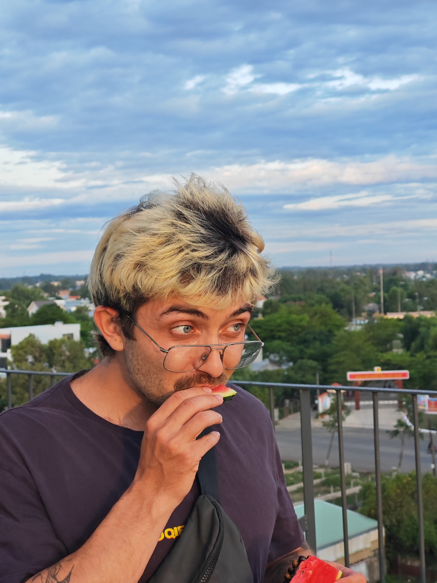 A man with blonde, spiky hair and glasses is eating a slice of watermelon outdoors. He is wearing a dark t-shirt and a black fanny pack. The background shows a cityscape with buildings and trees under a cloudy sky. The photo was taken in Đà Nẵng, Vietnam.