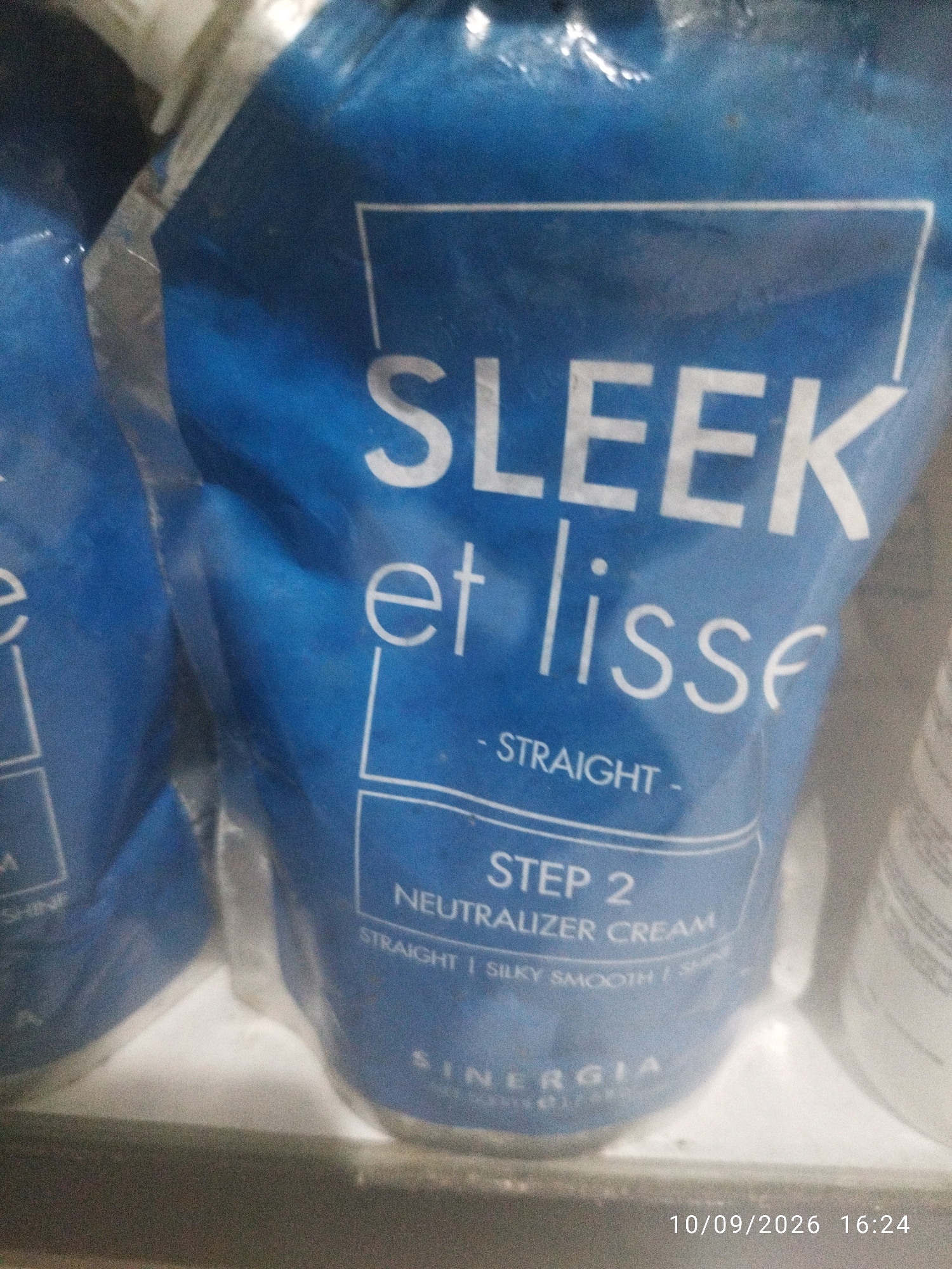 The image displays a close-up view of a blue pouch containing "SLEEK et lisse" hair straightening cream. The pouch is partially obscured by another similar pouch on the left. The text on the pouch indicates it is "STEP 2 NEUTRALIZER CREAM" and promises "STRAIGHT | SILKY SMOOTH | SHINE". The brand name "SINERGIA" is visible at the bottom. The pouch is placed on a dark, shelf-like surface. The background is blurred and out of focus, but appears to be indoors. A timestamp at the bottom right reads "10/09/2026 16:24". The image was taken in Mojokerto, Indonesia. There are no people or discernable interactions visible.
