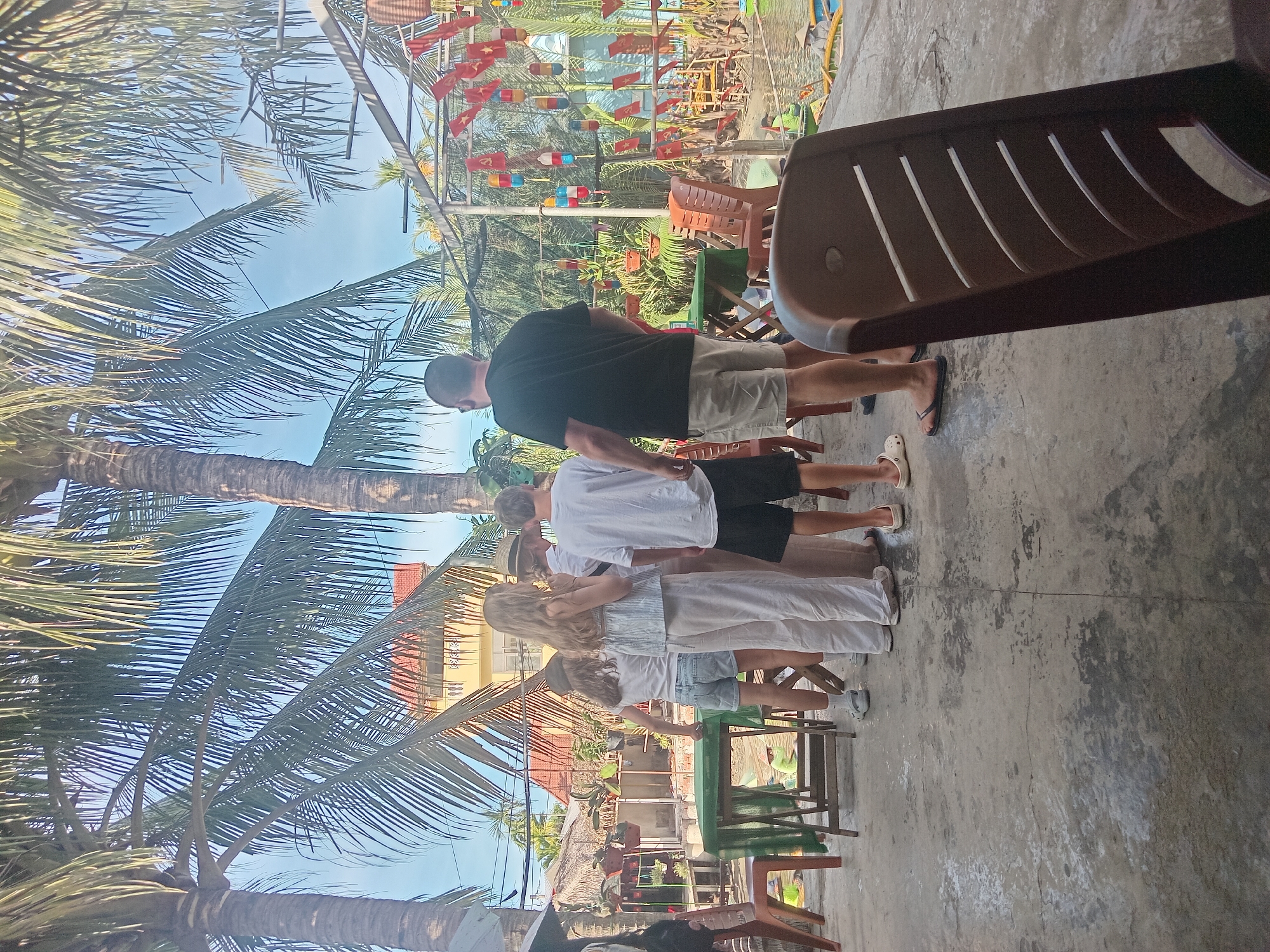 This media file depicts a group of people standing in an outdoor area, possibly a cafe or restaurant, with palm trees overhead. The scene appears to be set in Hoi An, Vietnam, as indicated by the presence of traditional Vietnamese flags and lanterns decorating the background. The people, a mix of adults and children, are casually dressed and seem to be engaged in a shared activity, perhaps a tour or a gathering.
The time of day appears to be daytime, with bright sunlight casting shadows, suggesting fair weather. The ground is concrete, and a dark brown plastic chair is prominently visible in the foreground. In the background, there are hints of other structures, possibly buildings with red roofs, and lush greenery. The overall atmosphere suggests a relaxed and tropical environment. There is no visible text in the image.