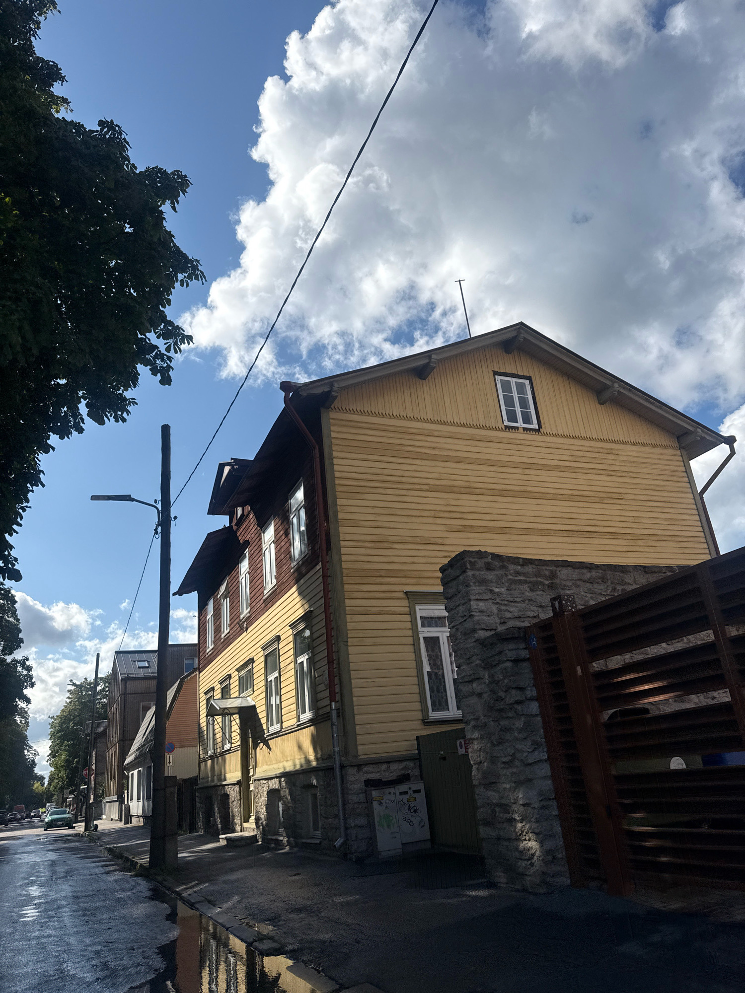 The image captures a street scene in Tallinn, Estonia, on a partly cloudy day. The viewpoint is low, looking up at buildings and the sky. On the right side of the frame, a two-story wooden house painted yellow with red trim is prominent. It features numerous white-framed windows. The lower portion of this building is a stone foundation. To the left of the yellow house, there is a dark wooden fence. Adjacent to the wooden house, a darker, multi-story building with red and yellow sections is visible.
In the foreground, a wet asphalt road reflects the sky and surrounding structures. There are puddles on the road, indicating recent rain. A black lamppost with a light fixture stands on the sidewalk. On the left side of the street, trees with green foliage overhang the scene. Further down the street, more buildings and a few vehicles can be seen. A green car is driving away from the camera on the wet road. The sky is a mix of bright blue and large, white cumulus clouds. A black power line runs diagonally across the upper portion of the image. The overall impression is a residential or mixed-use street with traditional wooden architecture, typical of some older parts of Tallinn.