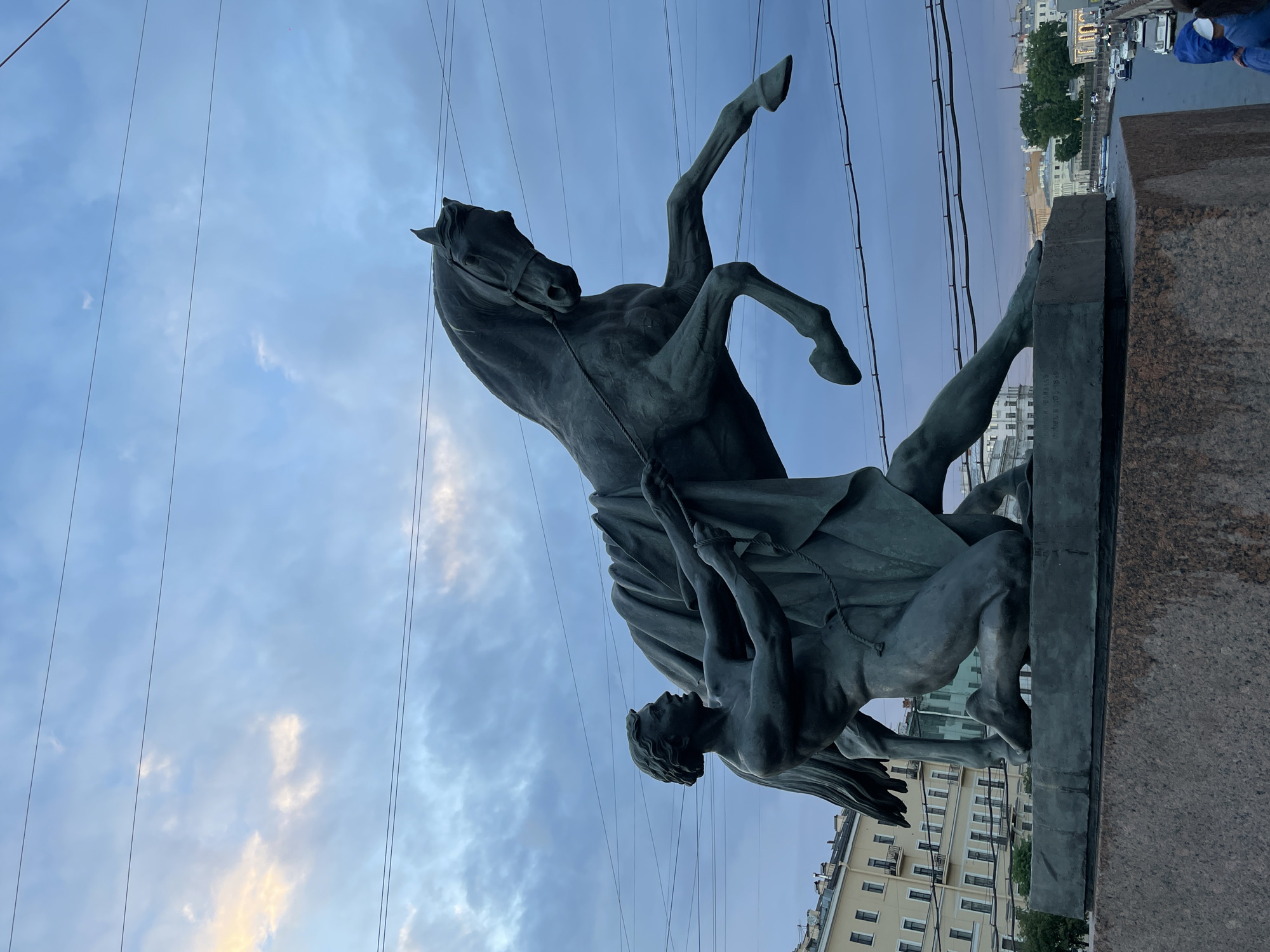The image shows a monumental bronze sculpture depicting a man subduing a rearing horse. The sculpture is situated on a granite pedestal, which is part of a bridge structure in Saint Petersburg, Russia. The man is on his knees, pulling on a rope attached to the horse's bridle, while a draped cloth covers the horse's body. The horse is captured in mid-air, with its front legs raised. In the background, there are buildings characteristic of Saint Petersburg architecture, and a river with some boats and cars on a bridge across it. The sky is cloudy with hues of blue and some light yellow. The scene appears to be taken during twilight. This sculpture is known as the "Horse Tamers" or "Riders" and is located on the Anichkov Bridge across the Fontanka River.