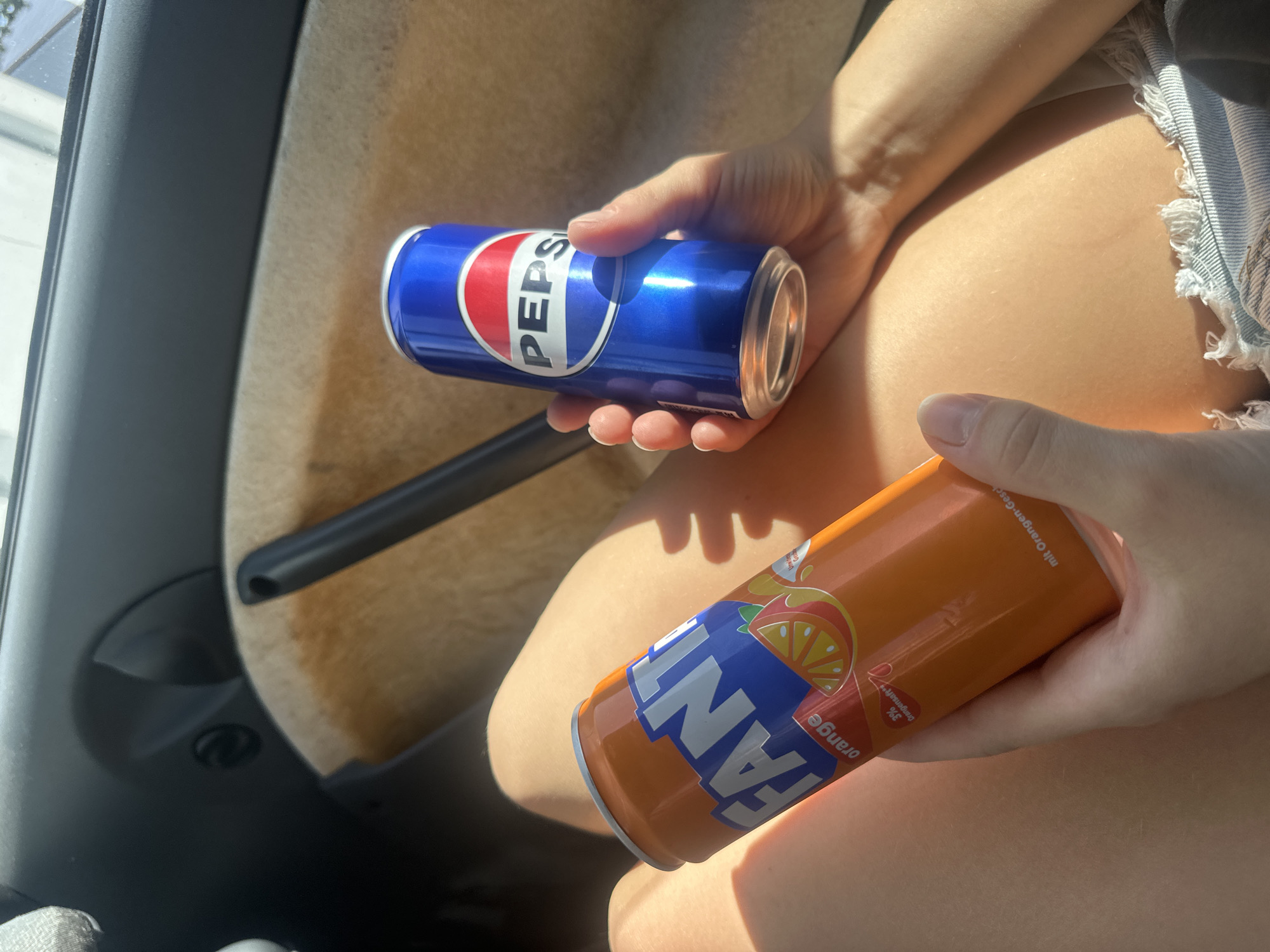 In the interior of a vehicle, two aluminum beverage cans are held by a person whose legs are visible. The can on the left is orange and displays the Fanta logo with an image of oranges. The can on the right is blue and red and features the Pepsi logo. The person is wearing denim shorts. The ambient light suggests it is daytime.