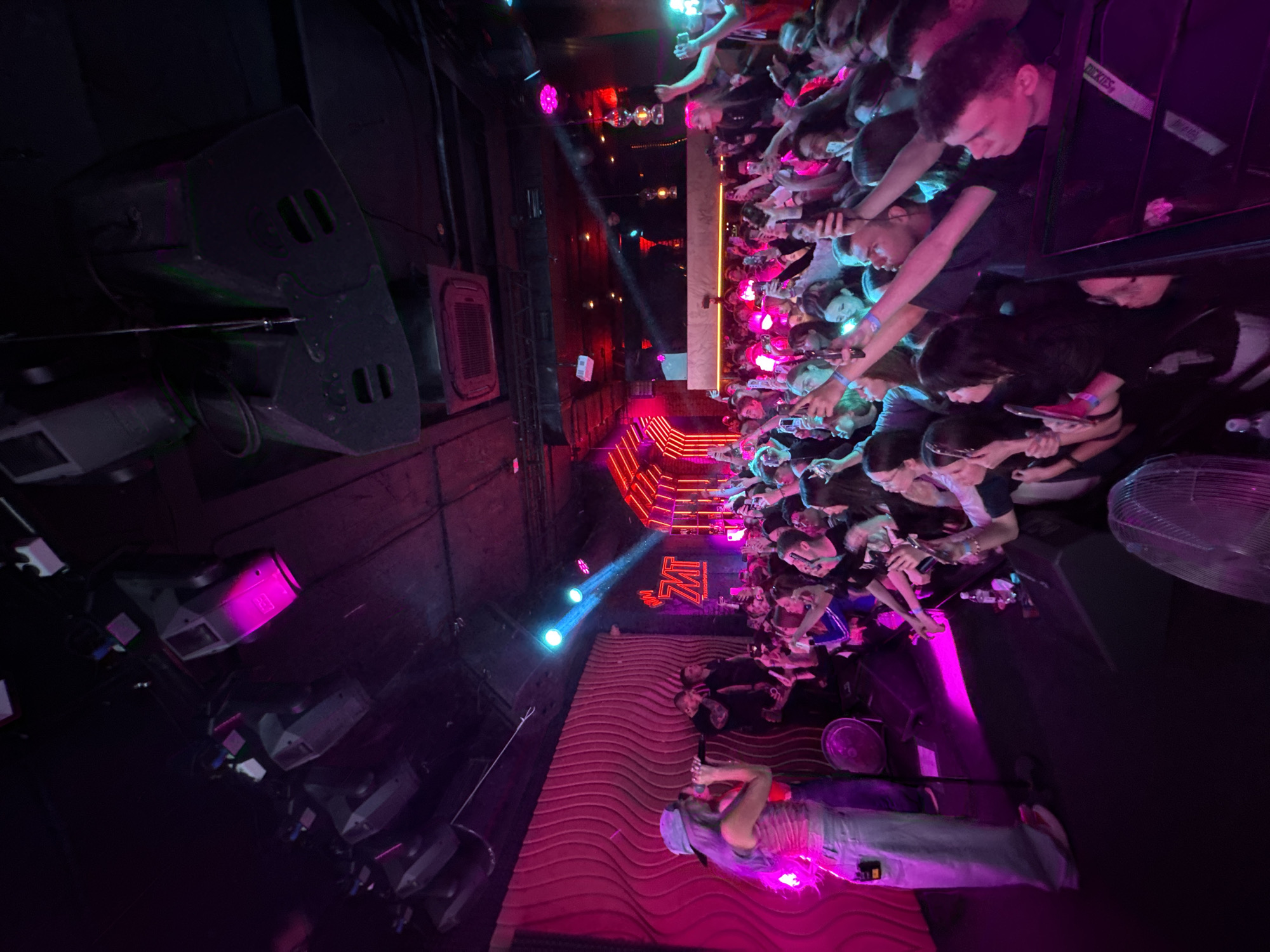 The image depicts a concert at the Velicano nightclub in Khabarovsk, Russia. A musician performs on a stage with a pink wave-patterned backdrop, illuminated by pink spotlights. The audience reaches towards the stage, many holding up phones to record. A DJ booth is visible at the edge of the crowd, and a red-lit staircase leads to an upper level where more audience members are present. Speakers are stacked on the left side of the image. The lighting is predominantly dark with pink and blue accents.