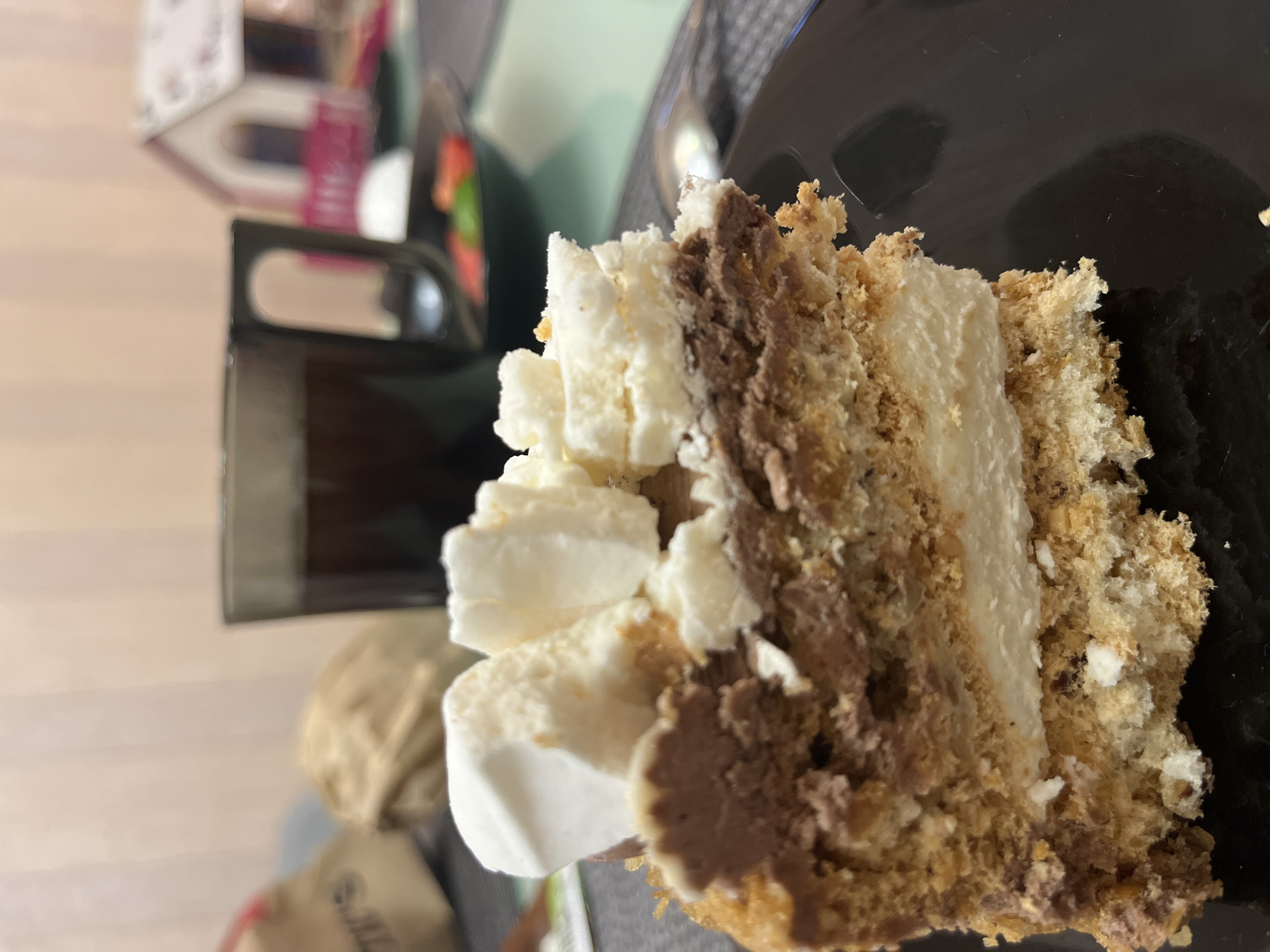 A close-up, eye-level shot shows a slice of layered cake on a black plate. The cake has a base of crumbly pastry, followed by a creamy white layer, then a chocolate layer, and topped with dollops of white whipped cream. In the background, out of focus, a dark mug of coffee sits on a table. To the right of the mug, a small, out-of-focus decorative house can be seen, resembling a dollhouse. Further to the right, a spoon is partially visible. The setting appears to be indoors, likely a home or cafe, with a blurred background suggesting a domestic environment. The image does not contain any specific indicators of the city of Sochi, Russia.