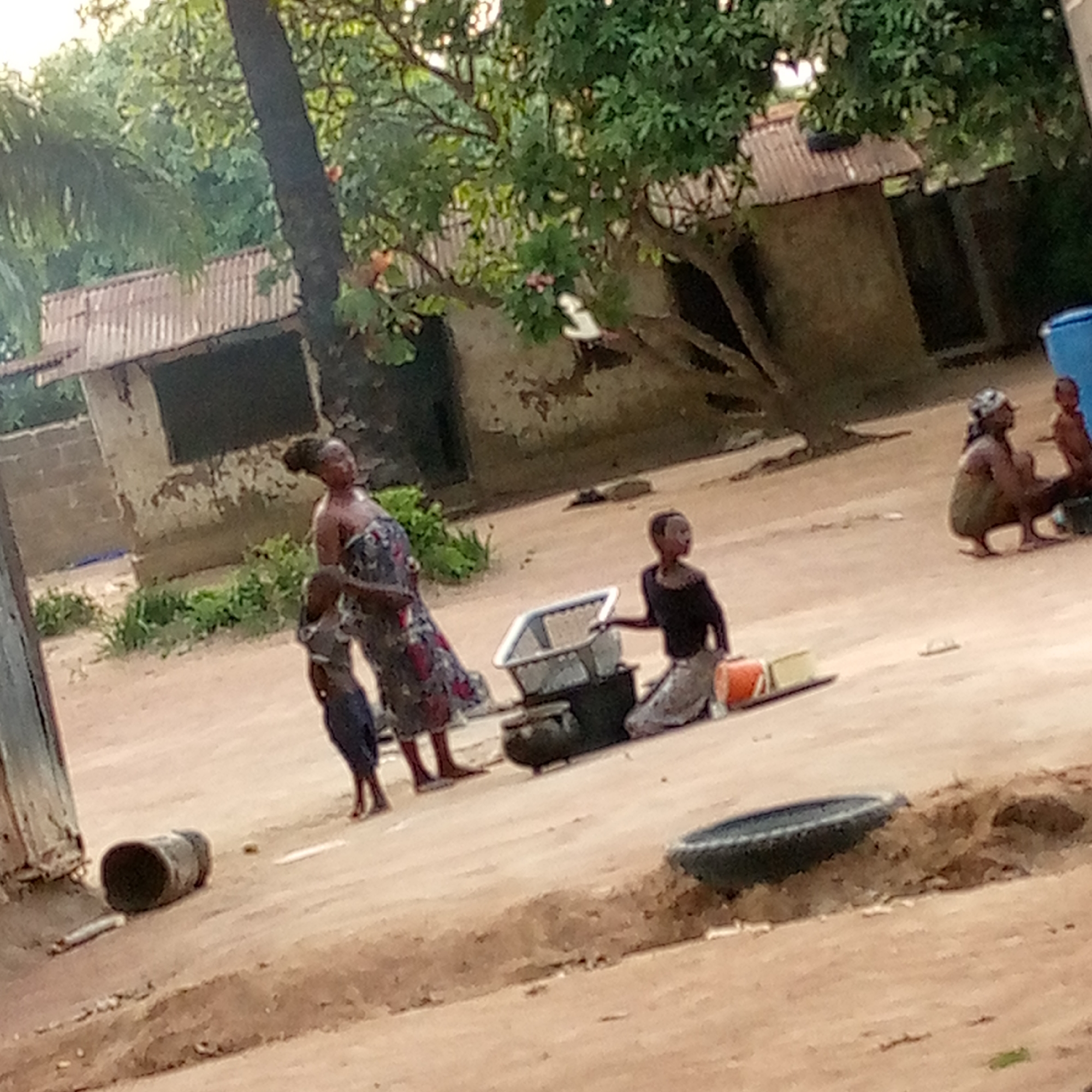 The image, taken in Mararaba, Nigeria, shows a group of people in an outdoor setting. A woman stands next to a child, while another child kneels near a cooking setup consisting of a metal container and a black charcoal stove. Other people are sitting in the background near a building. The surrounding environment includes simple buildings with corrugated metal roofs and trees. Scattered objects such as tires and containers are visible on the ground.