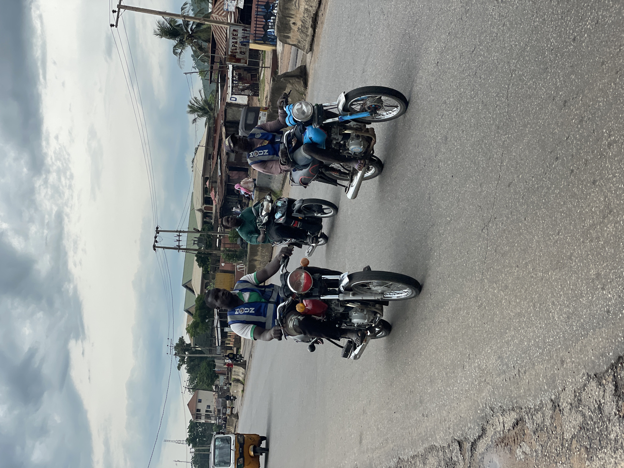 Three men ride motorcycles on a paved road in Lokoja, Nigeria. The man in the foreground on the red motorcycle wears a green and white shirt and a blue vest with "ZION" visible. The man in the middle on a black motorcycle wears a green shirt. The man on the right on a blue motorcycle wears a pink shirt and a blue vest with "ZION" visible. The road has patches of damage. In the background, buildings, palm trees, and power lines are visible under a cloudy sky. A yellow auto-rickshaw is also visible on the road.