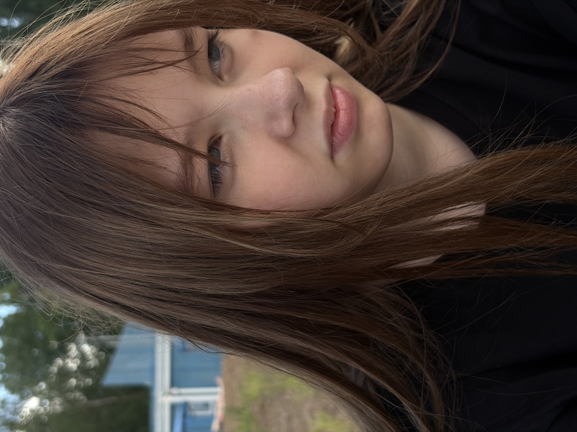 A close-up, medium shot captures the face of a young Caucasian female with long, light brown hair that falls across her left cheek and over her shoulder. She is wearing a black top. Her eyes are light blue and she is looking off to the right of the frame. She has fair skin with some freckles visible on her nose and cheeks. Her lips are slightly parted. The background is blurred, showing a blue wooden structure and some greenery, suggesting an outdoor setting. The overall lighting indicates it is daytime, possibly late afternoon due to the warm tones. The image appears to be taken in Chaikovsky, Russia.