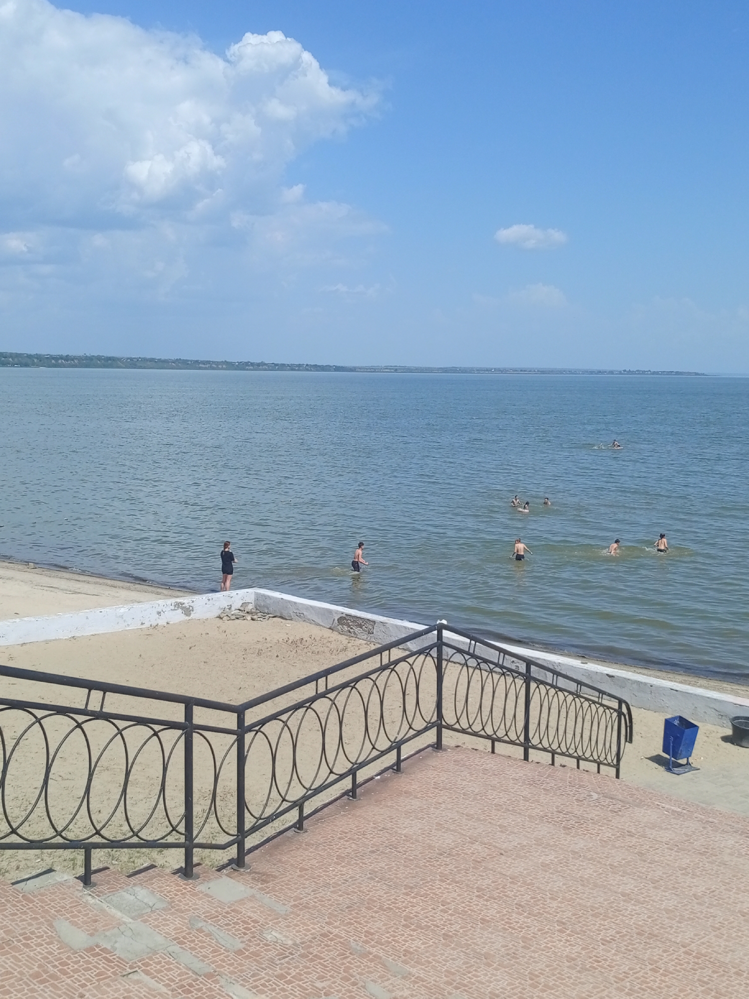The image shows a scene at a beach in an unknown city. In the background, the calm water of the sea or lake stretches to the horizon under a blue sky with scattered white clouds. Several people are swimming in the water at varying distances from the shore. On the shore, a person is standing near the water's edge. In the foreground, there's a sandy area with low white concrete retaining walls. A black metal railing with rounded shapes runs along the edge of a tiled area. A blue trash can is visible near the railing. The tiled area appears to be a walkway or steps leading to the beach.