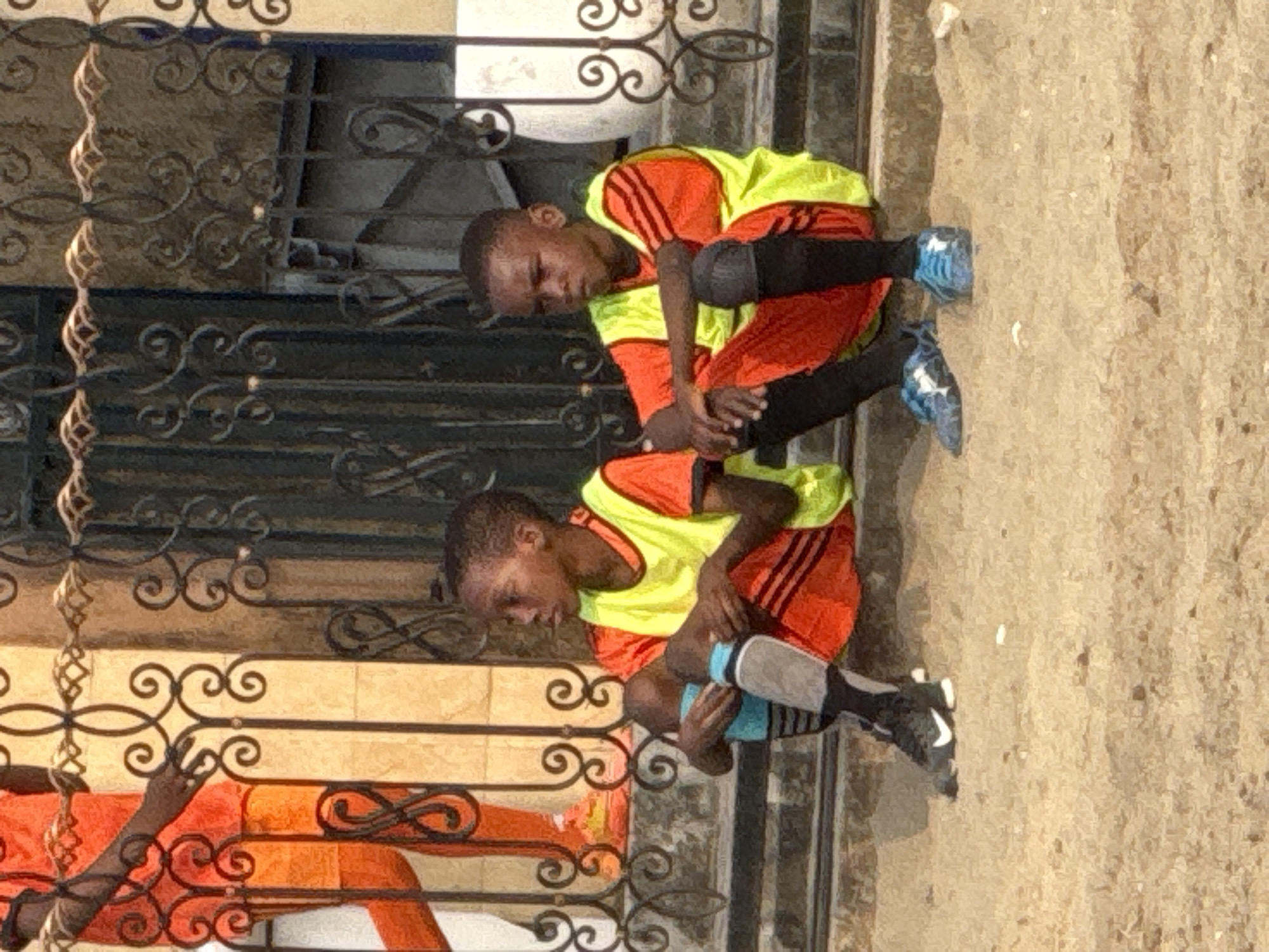 Two young boys, likely pre-teens, sit on a concrete step in an outdoor urban setting. They are wearing soccer uniforms consisting of orange jerseys with black stripes and yellow vests. The boy on the left has on black and gray striped socks with black shin guards and black Nike sneakers. The boy on the right wears black socks and blue patterned soccer cleats. Both boys are looking to their left. Behind them, a decorative black metal fence with intricate scrollwork separates them from a building with light-colored walls. The ground in front of them is sandy and appears to be a dirt path. The scene suggests a break during a soccer practice or game. The location is identified as being in the city of Ado, Nigeria.
