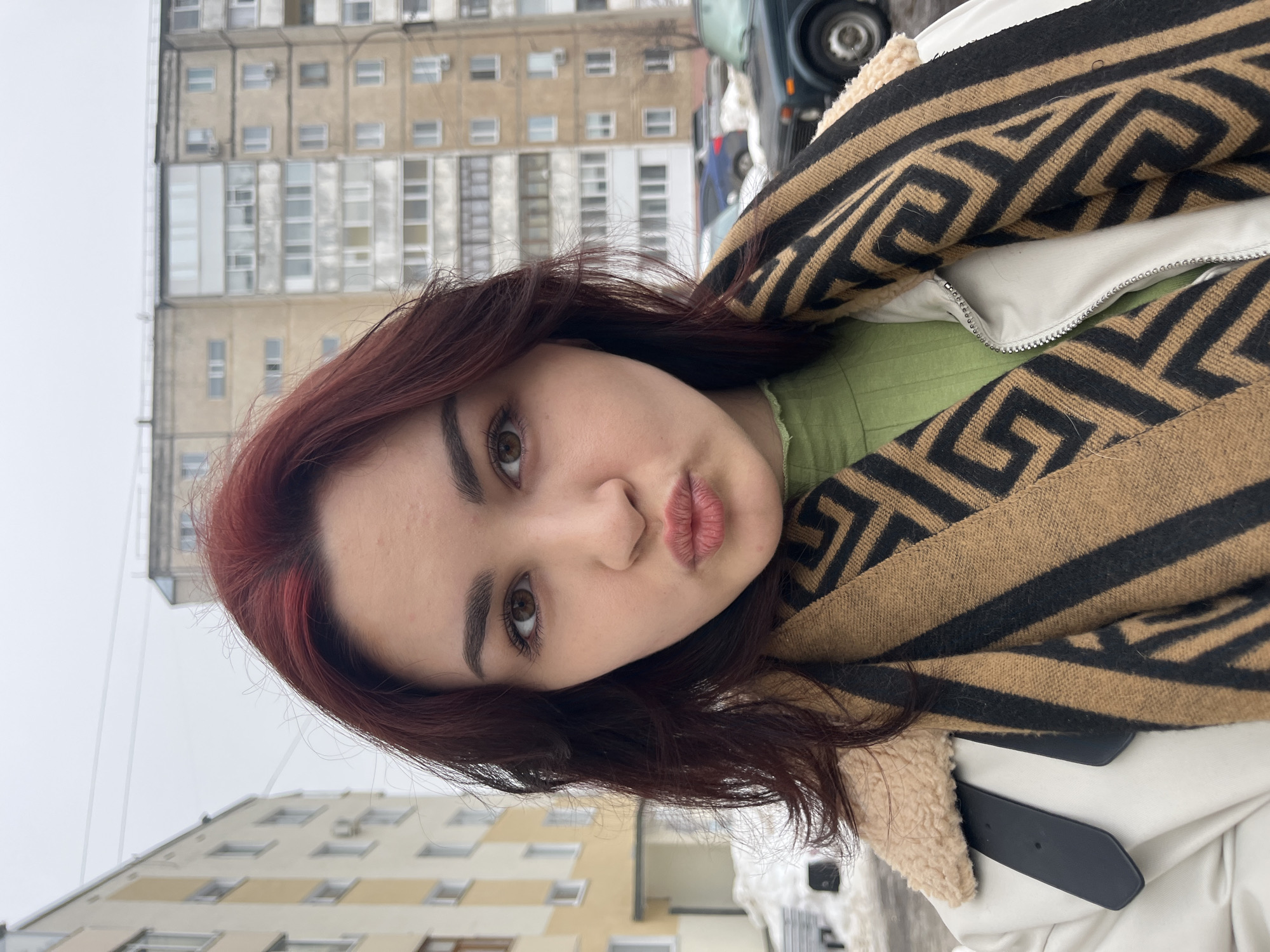 A young woman with dark red hair and brown eyes is centered in a selfie-style shot, making a pursed-lip or "kissing" expression. She wears a light-colored, possibly off-white or beige, jacket with a beige fleece-lined collar visible on the left shoulder, and a black and tan patterned scarf with a geometric design. Beneath the jacket, a light green top is visible. The background features residential buildings typical of Russian urban environments, specifically in Neftekamsk. To the immediate left is a yellow and white multi-story building, partially visible. To the right, a taller, multi-story concrete apartment building with numerous windows and some enclosed balconies extends upwards. Patches of snow are visible on the ground near the buildings and parked cars. The sky is overcast and uniformly light gray. A dark green car and other vehicles are partially visible parked on the right side of the frame.