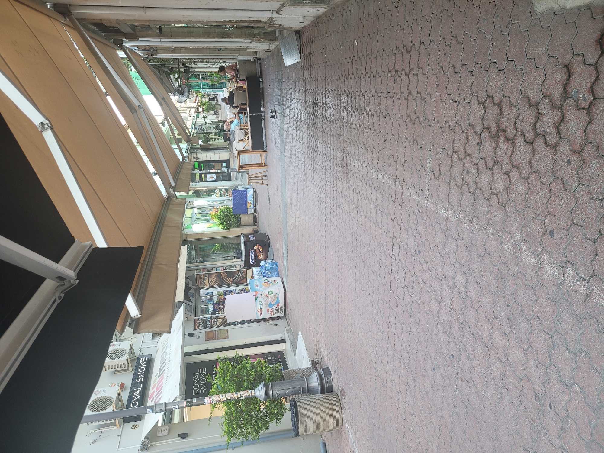 The image depicts a narrow pedestrian walkway lined with shops and awnings, typical of an urban commercial area. The ground is paved with red interlocking hexagonal bricks. On the left, a shop with a prominent "ROYAL SMOKE" sign is visible, featuring air conditioning units mounted on the facade. A lamppost with several stickers is positioned in front of this shop. Further down the walkway, there are more shops, some with display windows, and an ice cream freezer labeled "minimiers" and "Snickers". Several people are seated at tables outside a cafe or restaurant further down the walkway, engaging in conversation. The overall atmosphere suggests a pleasant, casual day in a city like Limassol, Cyprus, given the architectural style and the presence of outdoor seating areas.