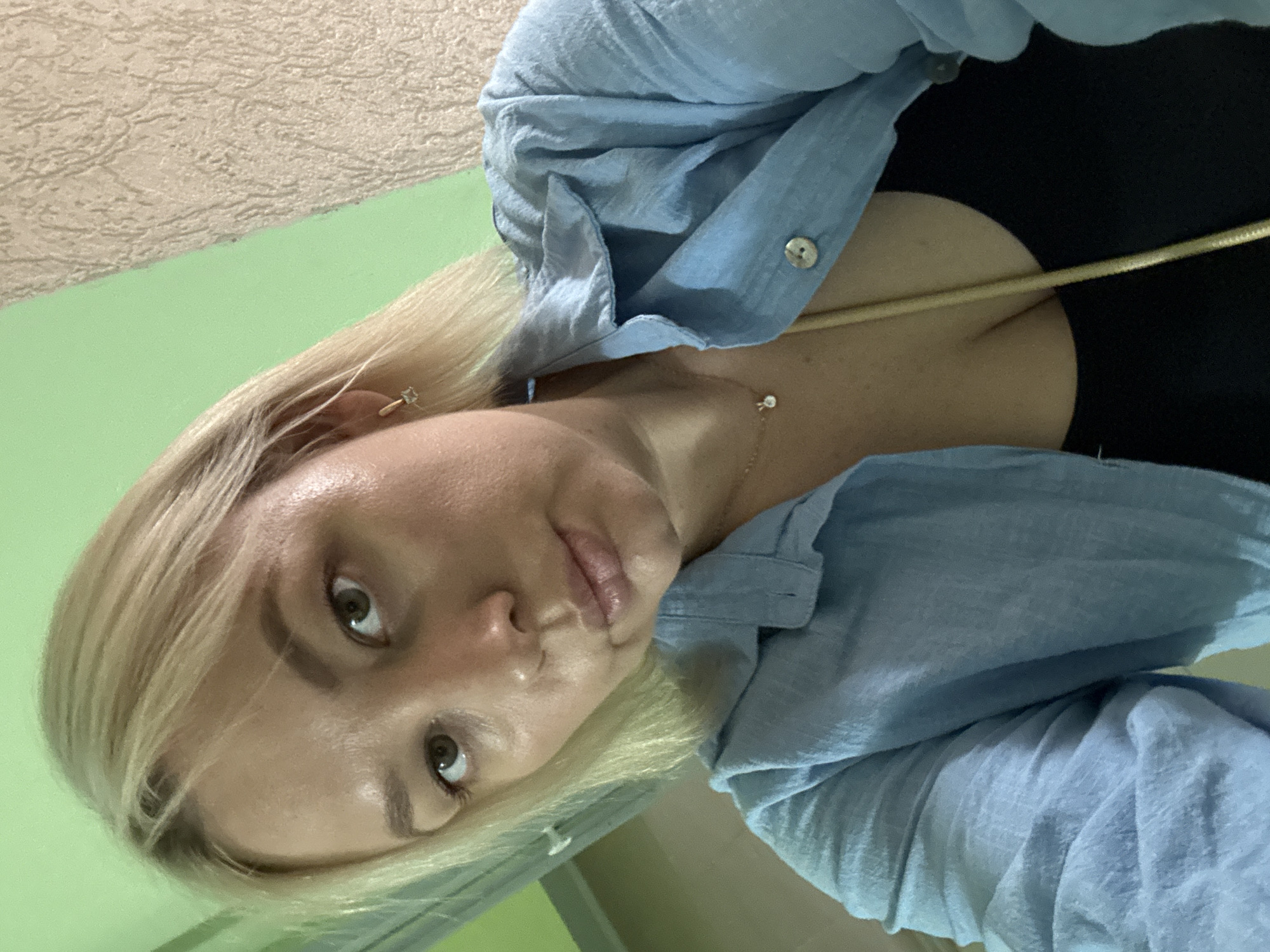 A woman with blonde hair and green eyes is looking at the camera. She is wearing a light blue button-down shirt over a black top, and a gold necklace. The background appears to be an indoor setting with light green walls. The image is a selfie taken from a slightly high angle. There are no specific identifiable landmarks within the image that would indicate a location in Chaikovsky, Russia.