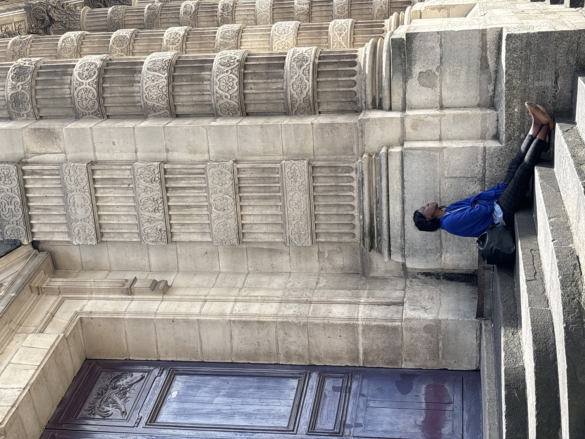 A woman of color is sitting on stone steps in front of a large, ornate building with columns. She is wearing a blue cardigan over a light-colored shirt and checkered pants. Her eyes are closed, and she appears to be resting or contemplating. A black backpack is beside her. The building's facade is made of stone and features intricate carvings. The scene is lit by natural sunlight, casting shadows on the steps. The architectural style of the building suggests it is a significant historical or religious structure in Paris, France.