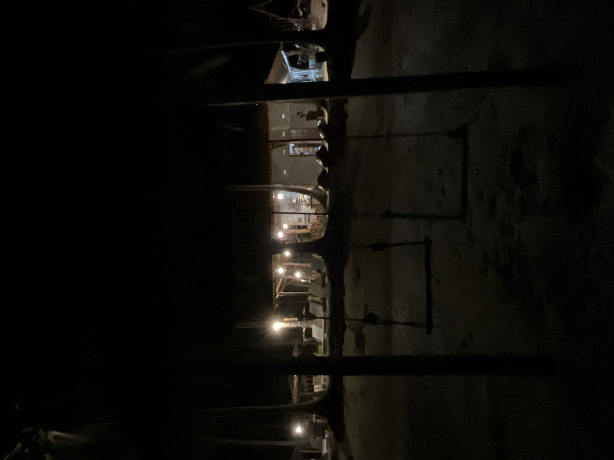 This is a nighttime photograph taken at a beach resort in Legonipah, Indonesia. In the foreground, there are two empty swings hanging from unseen structures, casting dark shadows on the sandy ground. Beyond the swings, illuminated by artificial lights, is a row of buildings that appear to be part of the resort. These buildings have open-air sections and are lit from within, revealing glimpses of furniture and possibly patrons. Palm trees are visible on either side of the scene, their silhouettes dark against the night sky. Lounge chairs are also visible in front of the resort buildings, suggesting a relaxation area. The overall atmosphere is tranquil and dimly lit.