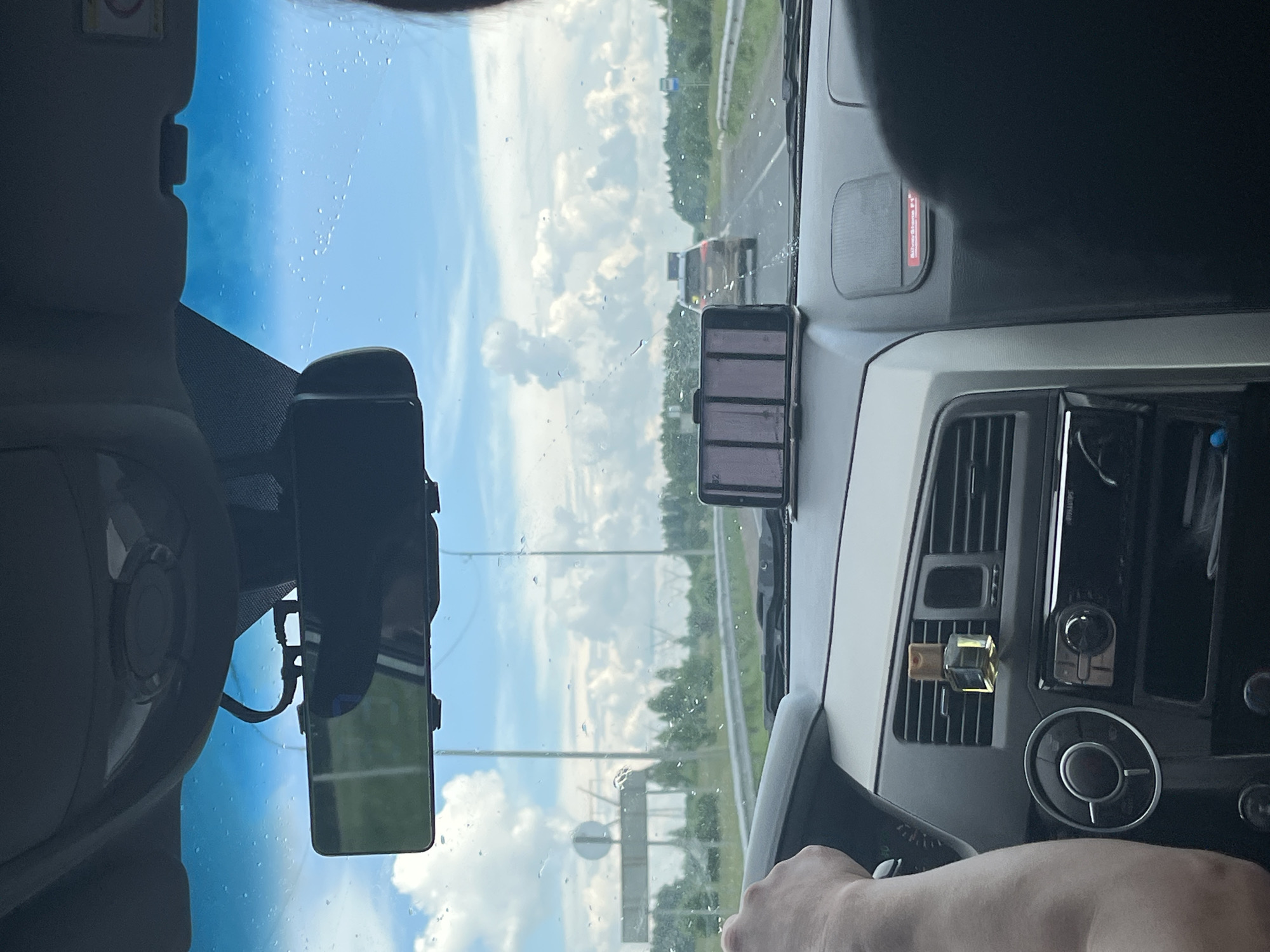 The image depicts the interior of a vehicle driving on a highway. The perspective is from the passenger seat, looking forward through the windshield. A person's left hand is visible on the steering wheel. The dashboard features a car air freshener, a car stereo, and a phone mounted on a holder displaying a pinkish-purple grid. Outside the vehicle, a highway stretches ahead with trees and a cloudy sky. In the distance, other vehicles and roadside signs are visible. The image appears to have been taken in Goldyrevskii, Russia, given the context provided.