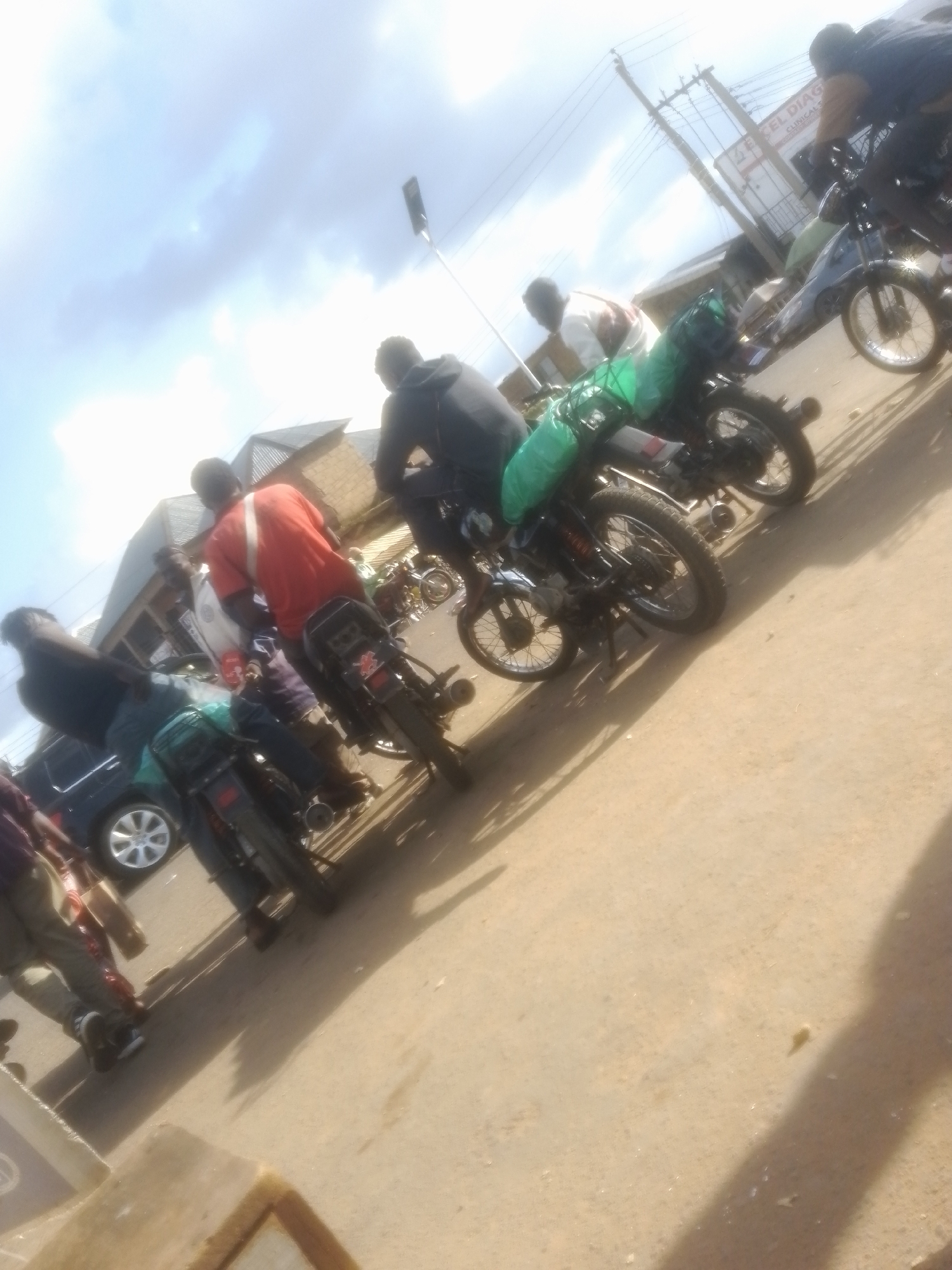 The image depicts a street scene in Gwong, Nigeria, with several people on and around motorcycles. The setting appears to be a bustling area, possibly a market or a transportation hub, given the presence of multiple motorcycles and a general sense of activity. The sky is visible in the upper portion of the image, with clouds suggesting it could be daytime, with varying light conditions.
The main subjects are people, some of whom are riding motorcycles, while others appear to be waiting or disembarking. The motorcycles themselves are prominent objects, with some carrying luggage or goods, indicated by green bags strapped to the back of some of them. A dark-colored car is also partially visible, suggesting that other forms of transportation are also present. The ground is paved, indicating a road or street.
The activity appears to be related to transportation, with people using motorcycles as a mode of transport. The general atmosphere seems busy and functional. There are no strong indicators of specific emotions, but the scene conveys a sense of everyday life and movement.
Visible text includes a sign in the background which appears to read "XEL DIAG" followed by "CLINICAL S." This suggests the presence of a diagnostic center or clinic nearby. The overall impression is of a lively street scene in an African urban or semi-urban environment.