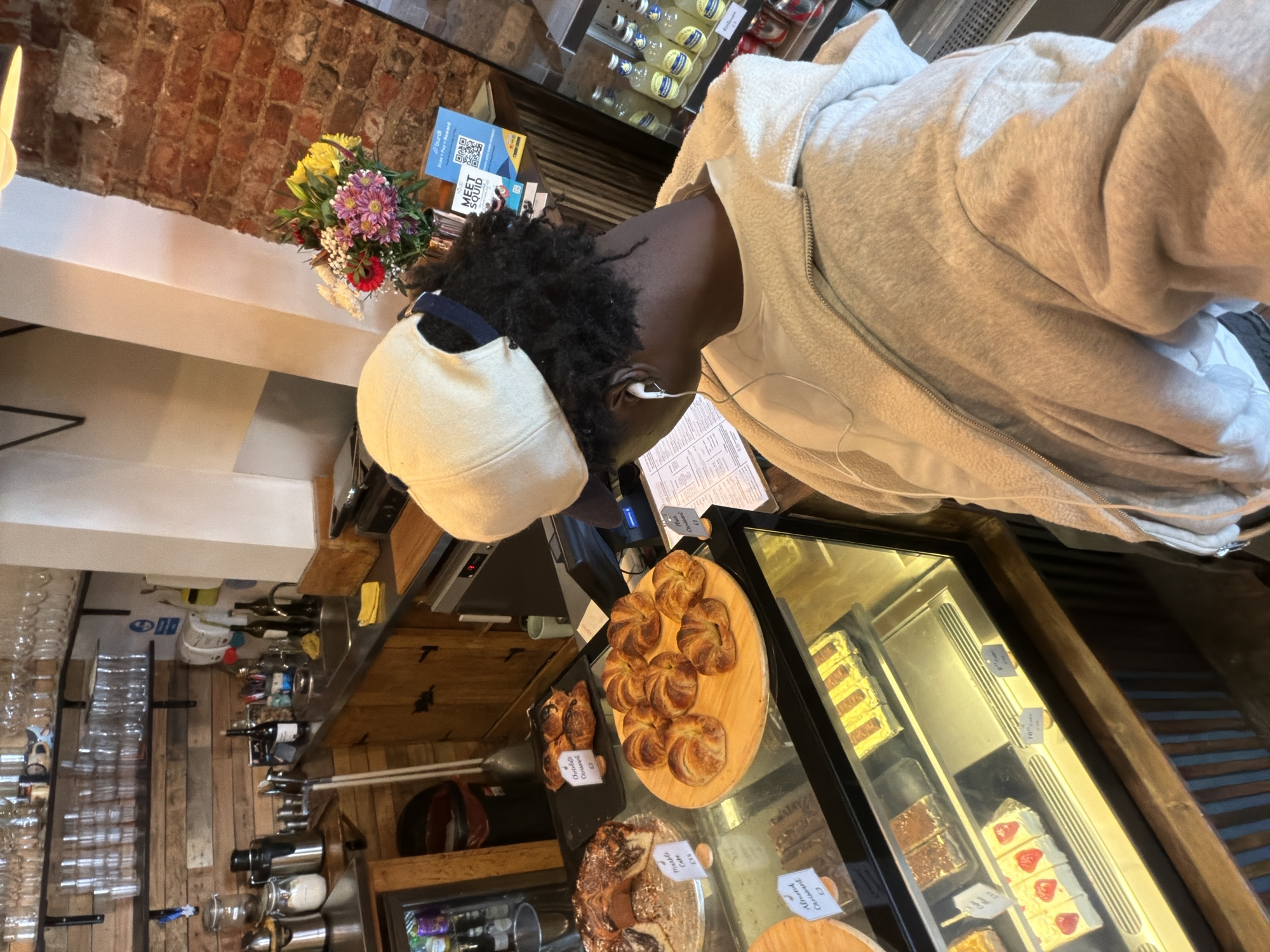 An overhead view captures a person inside a cozy cafe or bakery in London, United Kingdom, leaning over a display counter filled with tempting baked goods. The person, with dark skin and curly black hair, wears a light beige baseball cap, white wired earphones, and a light grey/beige zippered fleece jacket. They are looking down intently at the items in the glass display case.
On display are various pastries, including several golden-brown croissants arranged on a wooden platter, and other cakes and treats behind glass. Small white price tags are visible, reading "Almond Croissant £3", "Marble Cake £3.50", "Chocolate Croissant £3", and "Croissant £3". Behind the person, a refrigerated shelf holds multiple small bottles of beverages, likely lemonade or similar.
The cafe's rustic aesthetic is evident with a brick wall in the background, wooden shelving, and neatly stacked glassware. A colorful bouquet of yellow, purple, red, and white flowers adds a touch of vibrancy to the counter. A blue sign with a QR code and text "burdi", "Scan + Pay Reward", and "MEET SQUID" is also visible on the counter. The scene suggests a moment of contemplation before making a selection in a bustling cafe environment.