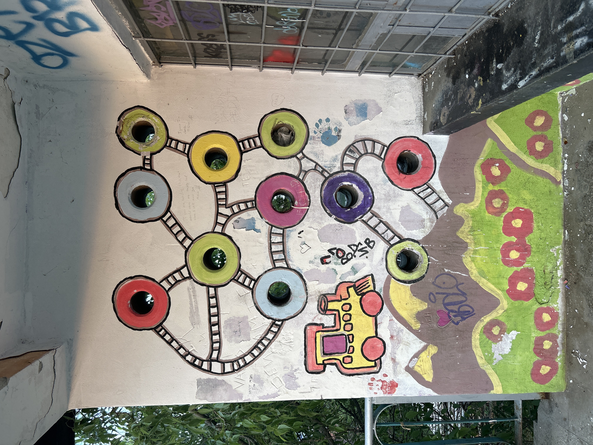 The image depicts an outdoor scene in Plovdiv, Bulgaria, featuring a wall adorned with colorful graffiti art. The wall is painted with interconnected circles of various colors, including yellow, green, red, purple, and gray, each with a central hole. Lines resembling ladders connect these circles. To the side, there is a drawing of a yellow vehicle, possibly a train or car, outlined in red and filled with red and purple details. Further along the wall, there's a section with brown and green paint, decorated with red flowers. Graffiti tags are scattered around the artwork, including the phrases "Sorry MOM," "I'm," and "If you are reading me happily," along with other scribbled text. A barred window is visible above the wall. Vegetation is visible at the bottom of the frame.