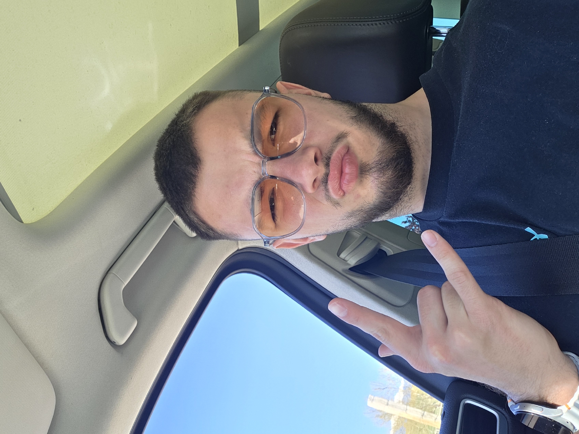 A man with a beard and tinted glasses is in a car with a sunroof on a sunny day. He is wearing a black shirt and giving a peace sign with his right hand. The location is not discernible from the image.