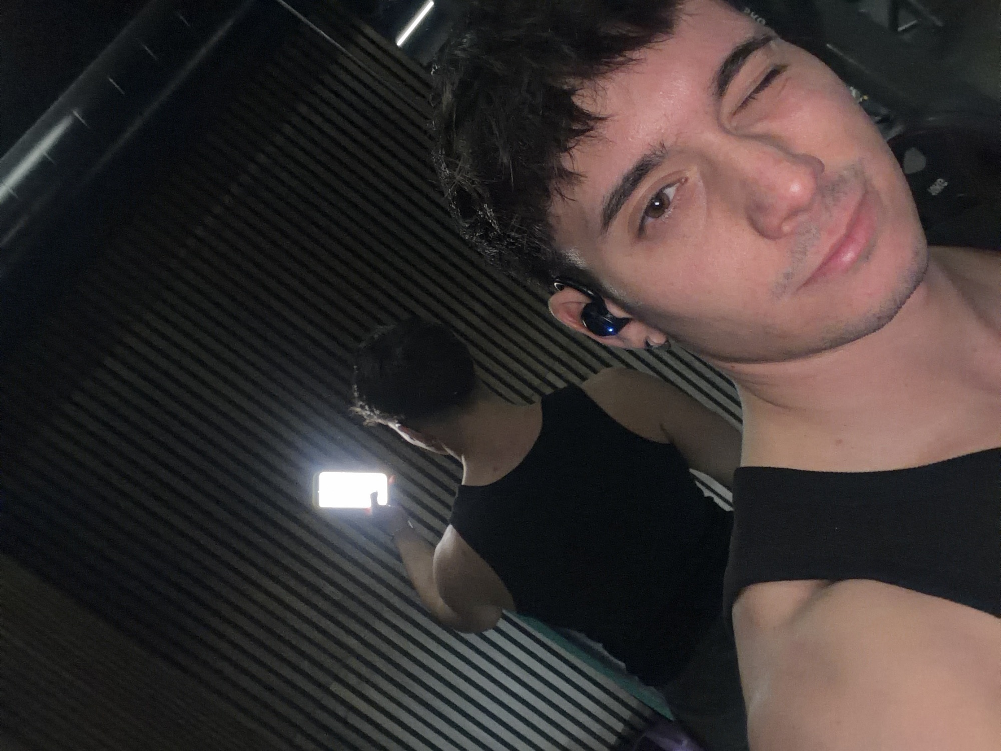 A selfie taken by a light-skinned male with brown hair and wearing a blue earbud in his left ear, winking his left eye. The man is wearing a black tank top. Reflected in a wall of vertical slats is another person wearing a black tank top and holding a brightly lit smartphone horizontally. The exact location in Sofia, Bulgaria cannot be determined based on the image.