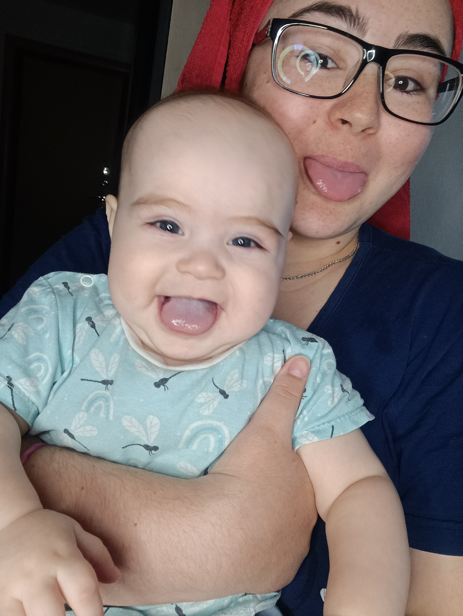 A medium shot depicts a woman holding a baby. The woman is wearing glasses and has a red towel wrapped around her head. She is sticking her tongue out. The baby, wearing a light blue shirt with a dragonfly and rainbow pattern, is also sticking its tongue out and smiling. The woman is wearing a dark blue t-shirt and a thin silver chain. The background shows a dark doorway and a gray wall. The location is in Noviy, Russia.