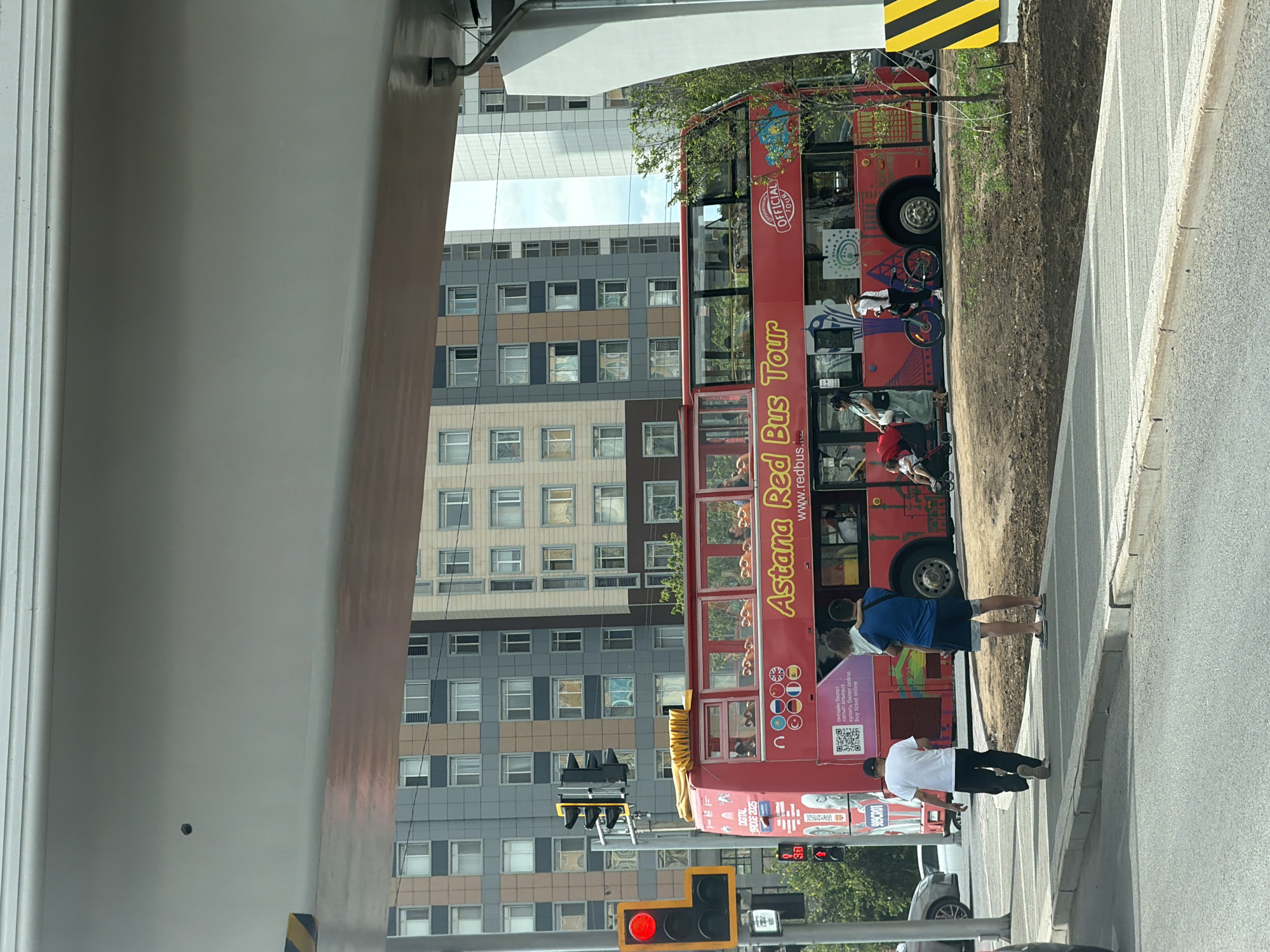 This image captures a street scene in Astana, Kazakhstan. The central focus is a bright red double-decker bus, branded as the "Astana Red Bus Tour." The bus is parked on the side of a street, with a sidewalk and a grassy embankment beside it.
Several people are visible near the bus. A person in a blue shirt and shorts is standing on the sidewalk, holding a child. Another person in a white t-shirt and black pants is walking away from the bus. The bus itself has its windows open, and some passengers can be seen inside. The bus displays promotional text in what appears to be Kazakh and English, including "Astana Red Bus Tour" and "buy ticket online."
In the background, a large, modern building complex with many windows rises up, suggesting an urban environment. Traffic lights are visible, one of which is red, indicating that vehicles may be stopped. The weather appears to be fair, with daylight suggesting it's daytime. The overall impression is of a typical urban setting where a tourist bus is operating. The presence of multiple languages on the bus suggests it caters to a diverse clientele.