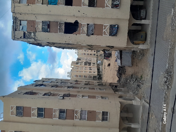 The image displays a desolate urban scene in the city of Khan Yunis, Palestinian Territory, dominated by extensively damaged multi-story residential buildings under a partially cloudy blue sky. Two prominent beige-colored apartment blocks with brown window accents flank a central open area, with additional damaged structures visible in the background.
The building on the left, approximately seven stories tall, exhibits visible damage including broken windows, some covered with opaque materials, and pitting across its facade. Its ground level features a series of open, arched passages. The building on the right, also multi-story, shows more severe destruction, with significant portions of its upper facade missing and large, dark apertures where windows once were. Multiple windows are either blown out or covered with blue tarps and fabric. Extensive pockmarking and spalling are evident across its entire visible exterior. Beneath this building, within an arched ground-floor section, one vehicle is largely covered by a yellow tarp, and another lighter-colored vehicle is partially visible.
The central area between the two foreground buildings is an unpaved, dirt and sand-covered expanse littered with heavy debris, including concrete fragments, twisted metal, and other rubble. A small, makeshift structure, covered with a white tarp, stands amidst this wreckage. Further into the background, a cluster of similarly damaged residential buildings can be seen, indicating widespread destruction. The immediate foreground reveals a dark, paved surface, possibly a street or walkway, with scattered small debris and two horizontal dark lines, consistent with overhead wires or cables. No individuals or active elements are discernible within the frame. The image is watermarked "Galaxy A14" in the lower left corner.