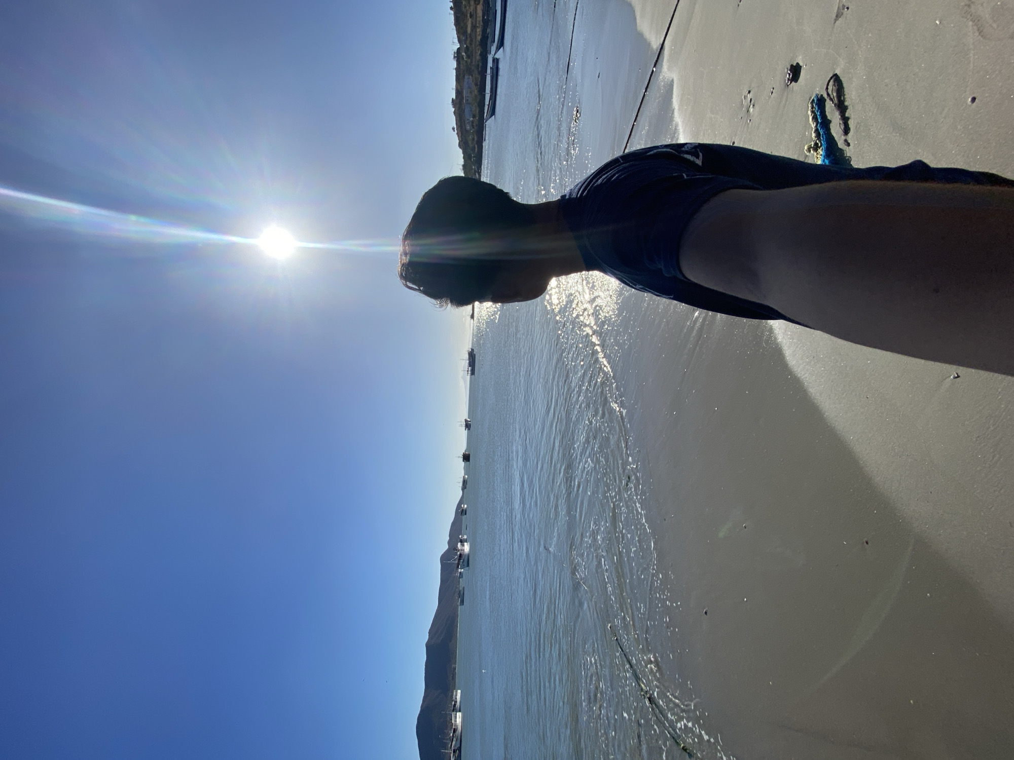 The photo shows a person at Playa La Galera in Juan Griego, Venezuela. The bright sun is in the background over the sea. Several boats are visible in the water. Part of the person's arm and shoulder are visible, and they appear to be wearing a dark t-shirt. The sandy beach and the water's edge are in the foreground.