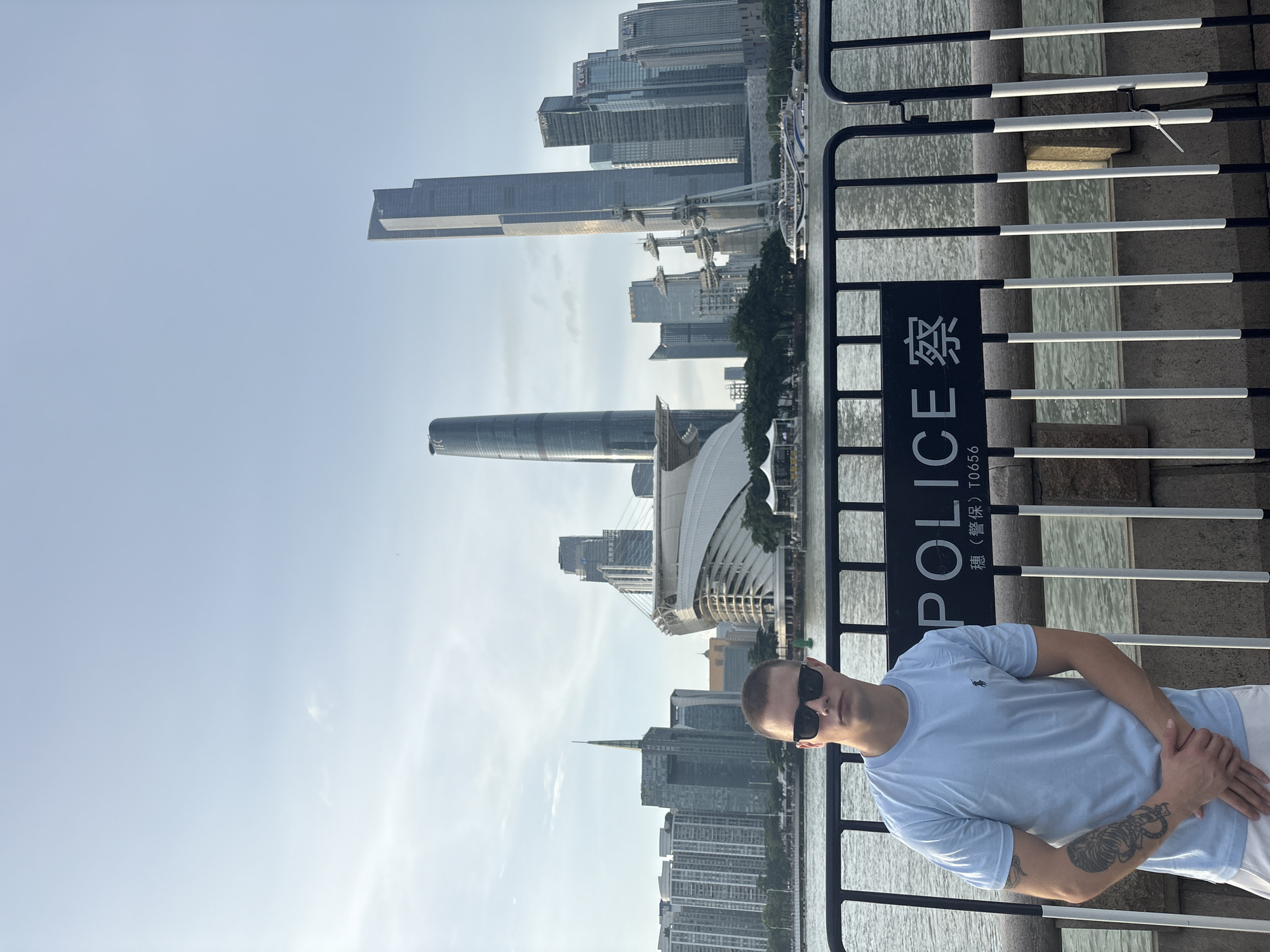 A young man stands in front of a metal barrier and a sign that reads "POLICE" in English and Chinese characters. He is wearing a light blue t-shirt, white shorts, and sunglasses. The background features a city skyline with numerous tall buildings and a body of water. The scene appears to be a waterfront promenade in the Haizhu District of China, with the Pearl River visible. A boat is seen on the water, and trees and other structures are present along the embankment. The sky is partly cloudy.
