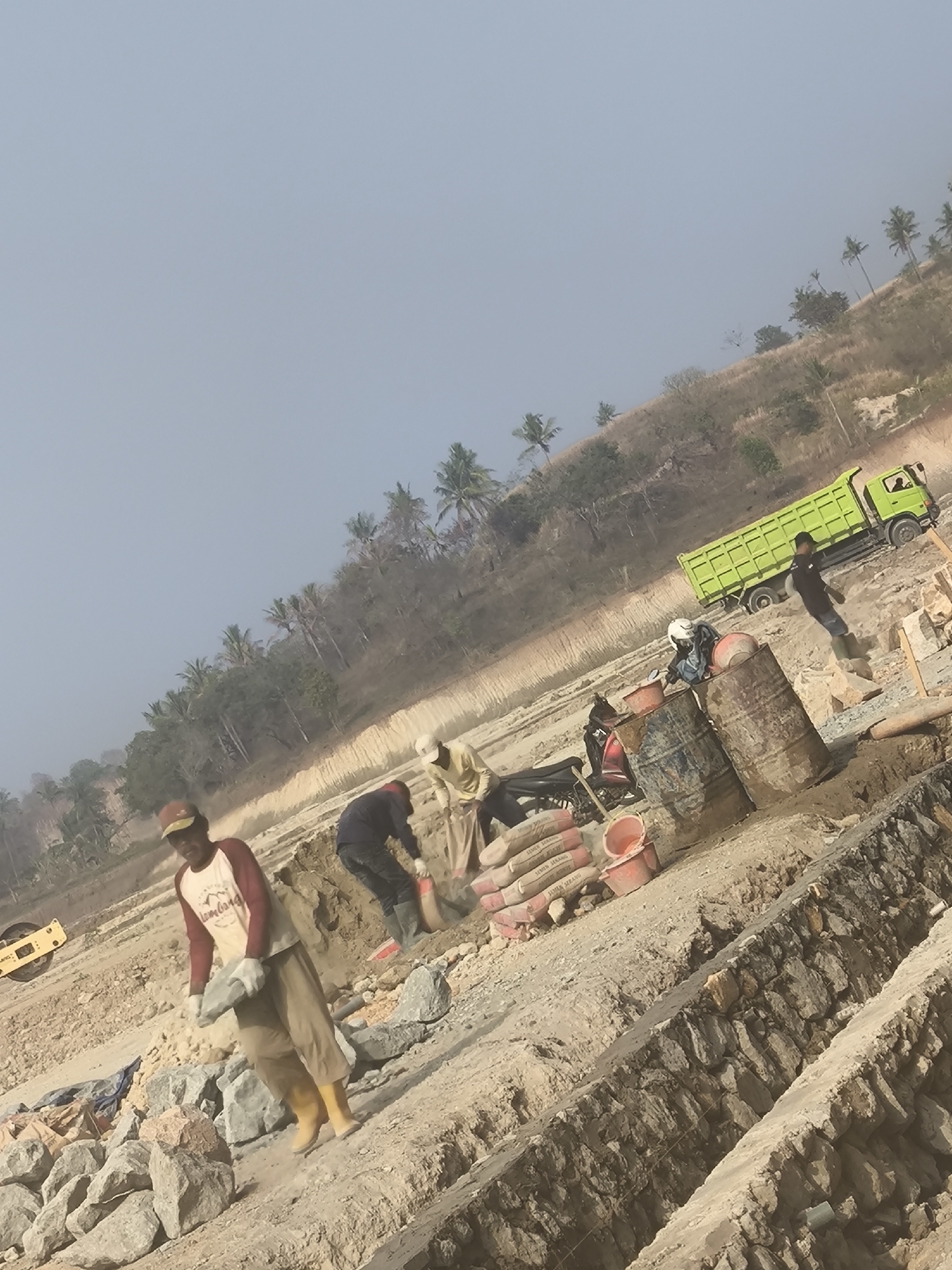In Cilegon, Indonesia, a construction site is depicted with several individuals engaged in manual labor. The scene is set on a sloping terrain with scattered vegetation, including palm trees, and a clear, pale sky. In the foreground, a construction worker wearing a red and white shirt, tan pants, and yellow boots carries a large flat stone. To his right, another worker in a dark shirt and green boots is partially obscured, appearing to be shoveling material. Further in the midground, a person wearing a white helmet is seated on a motorcycle, with two large metal barrels and red buckets nearby. A bright green dump truck is positioned on a dirt path in the background, facing left, with a person standing near its rear. The ground is a mixture of dirt, rocks, and construction materials, including several bags of cement. The overall impression is one of active construction work in a semi-arid environment.