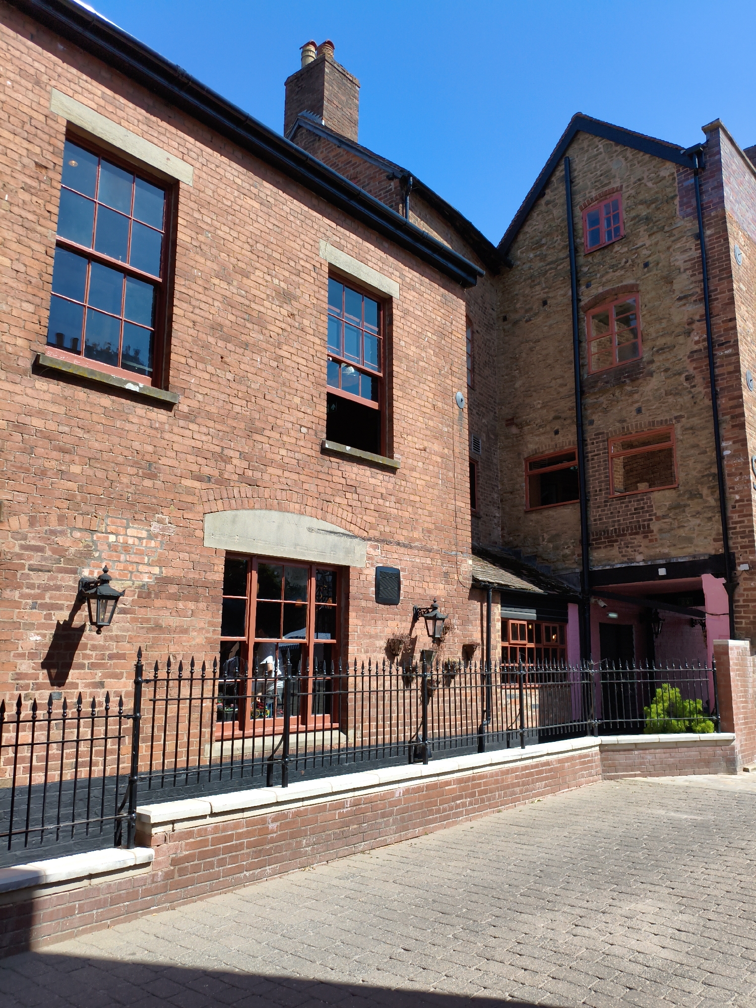 The image shows a courtyard area in front of a brick building with multiple windows and a chimney. The building has red brick walls and red-framed windows, some of which are divided into multiple panes. There is a black metal fence running along the front of the building, separating it from a paved courtyard. The sky is clear and blue. The building appears to be a historic structure, possibly a pub or restaurant, given the style of the windows and the presence of outdoor seating areas visible through the windows. The context provided suggests this location is in Ludlow, United Kingdom.